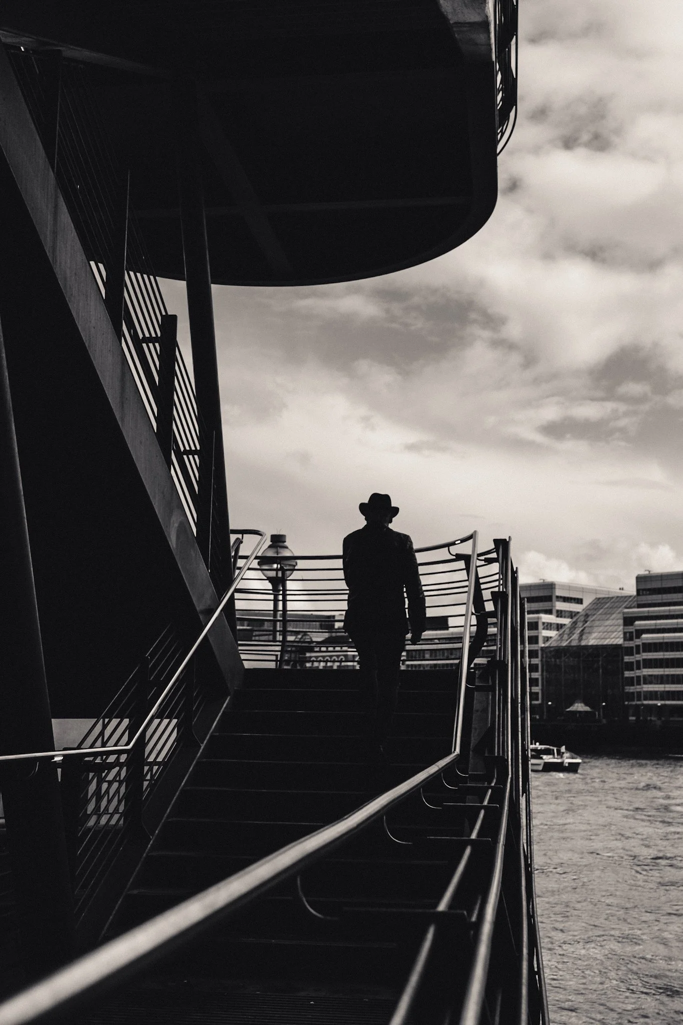 A person wearing a hat standing on an outdoor staircase next to a body of water, with city buildings in the background and clouds in the sky.