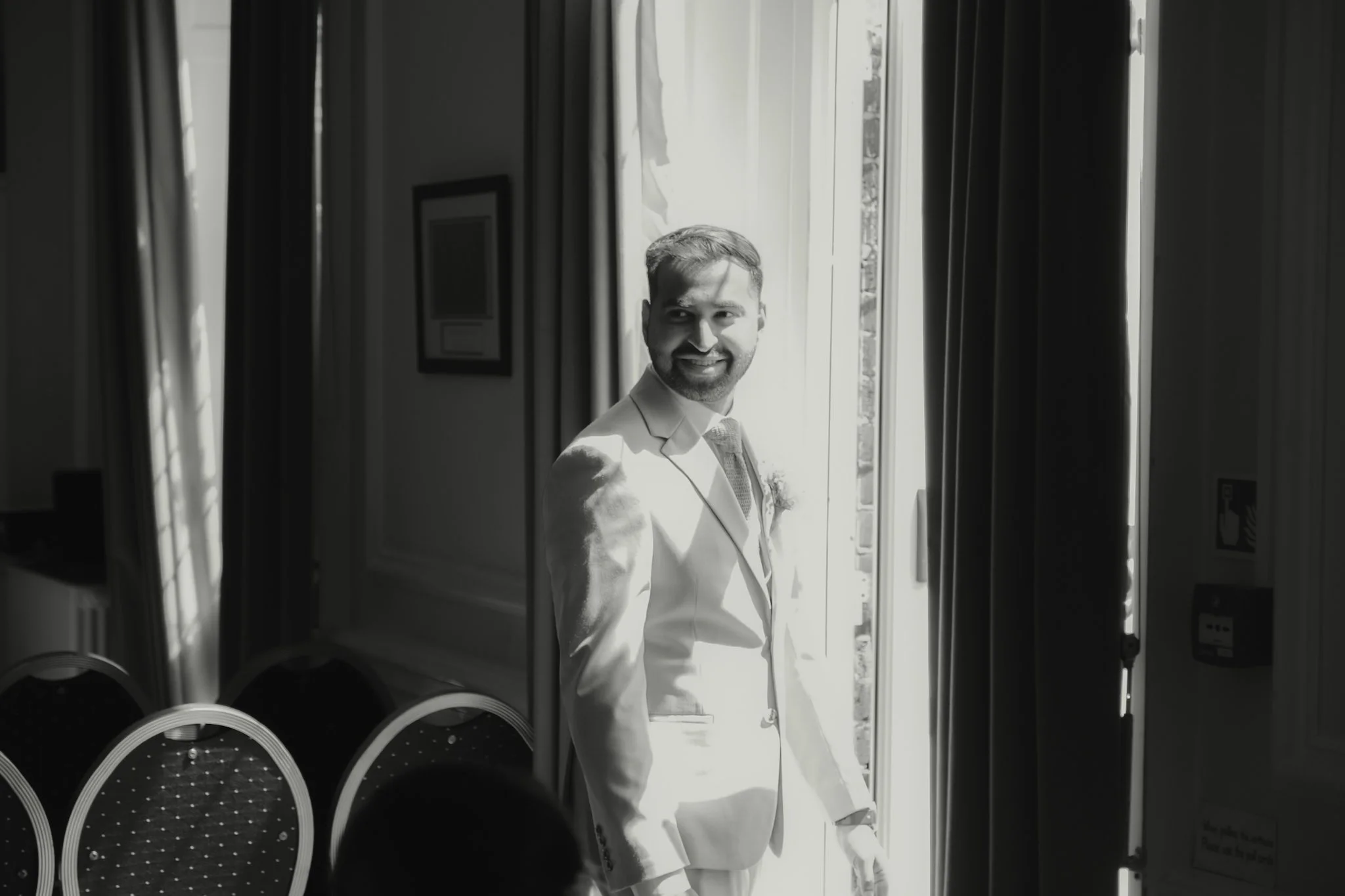 A man dressed in a white suit, smiling, standing by a door with sunlight coming in, in a room with black and white chairs.