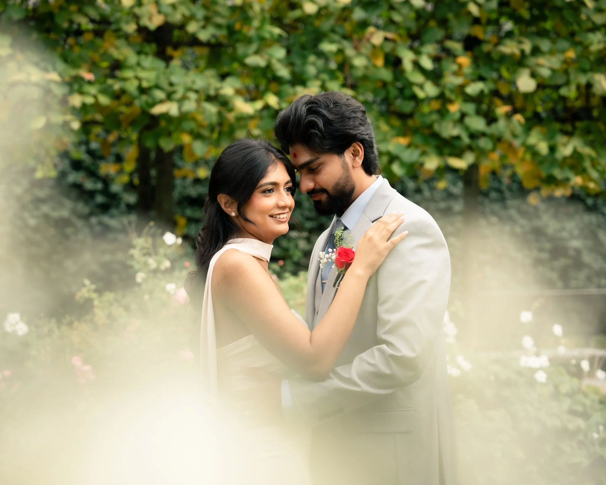 A couple dressed in wedding attire embracing outdoors with a lush green background.