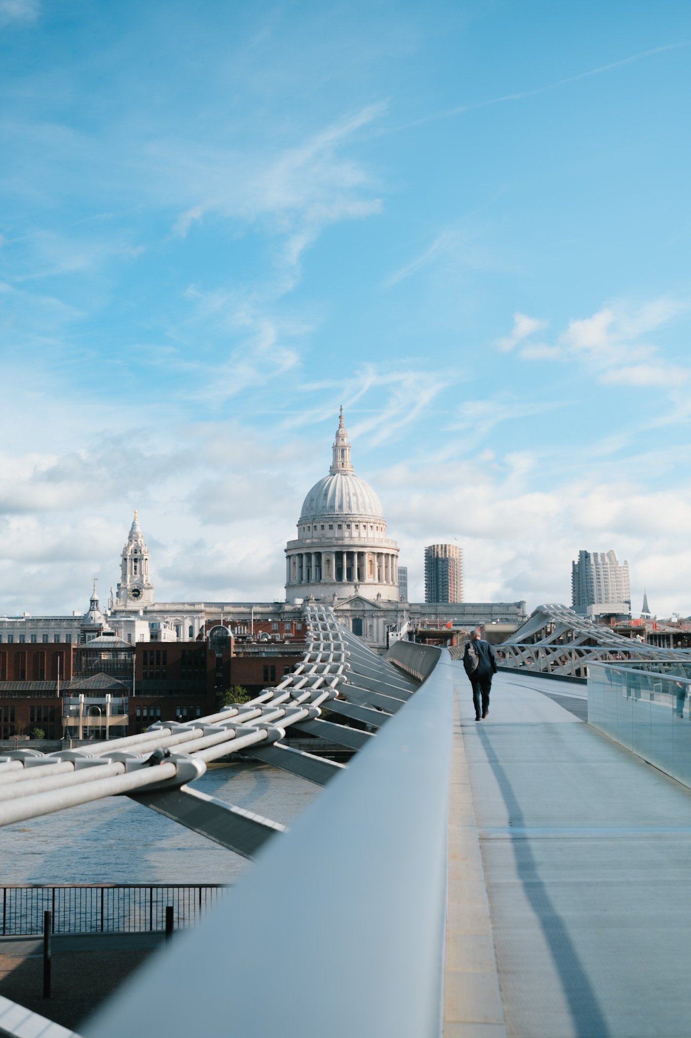 Cityscape featuring St. Paul's Cathedral in London, viewed from the Millennium Bridge with pedestrians, modern skyscrapers, and a partly cloudy sky.