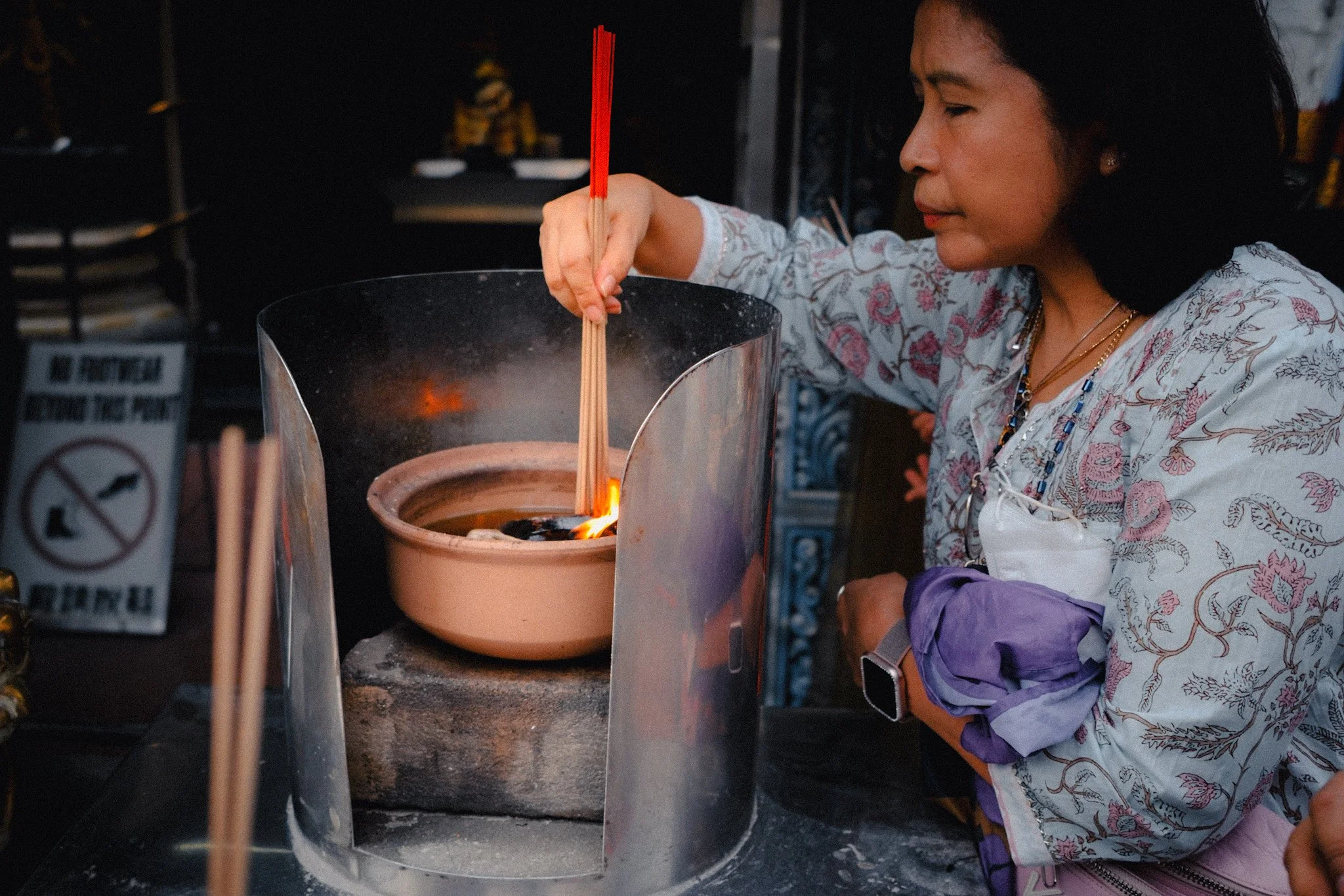 A woman wearing a floral blouse and purple apron is cooking with a small stove, stirring contents in a ceramic pot over an open flame. The scene appears to be in a food stall or outdoor kitchen.