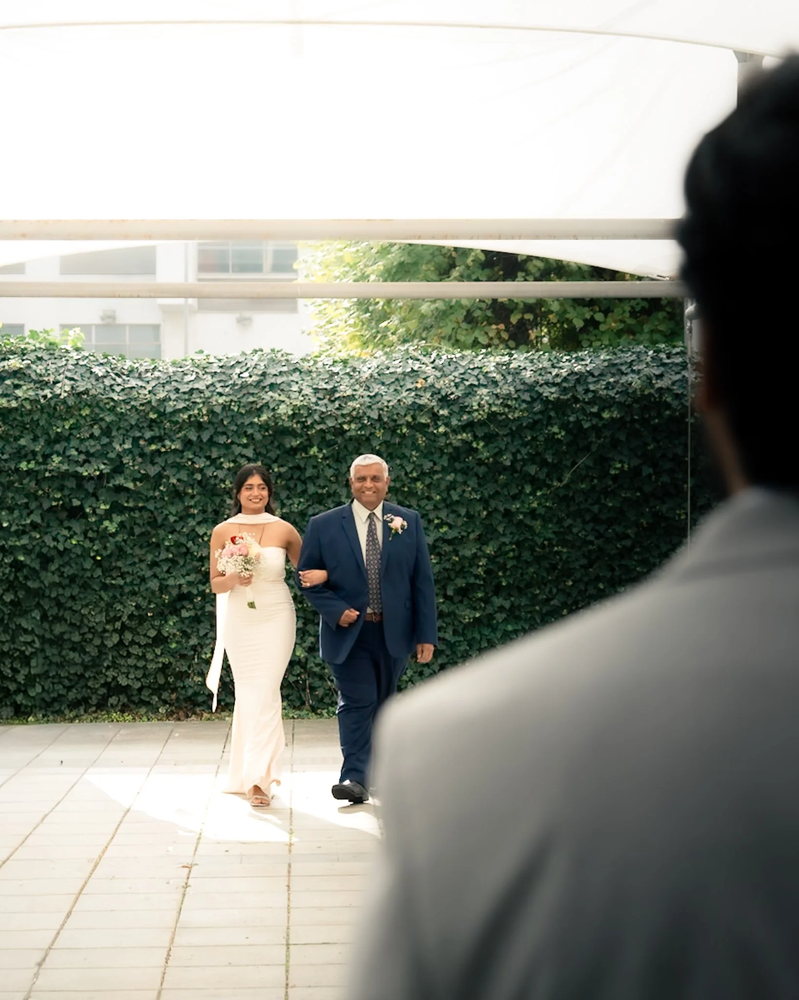 A couple walking outside in formal attire, the woman in a white gown holding a bouquet, and the man in a dark suit with a boutonniere, both smiling.