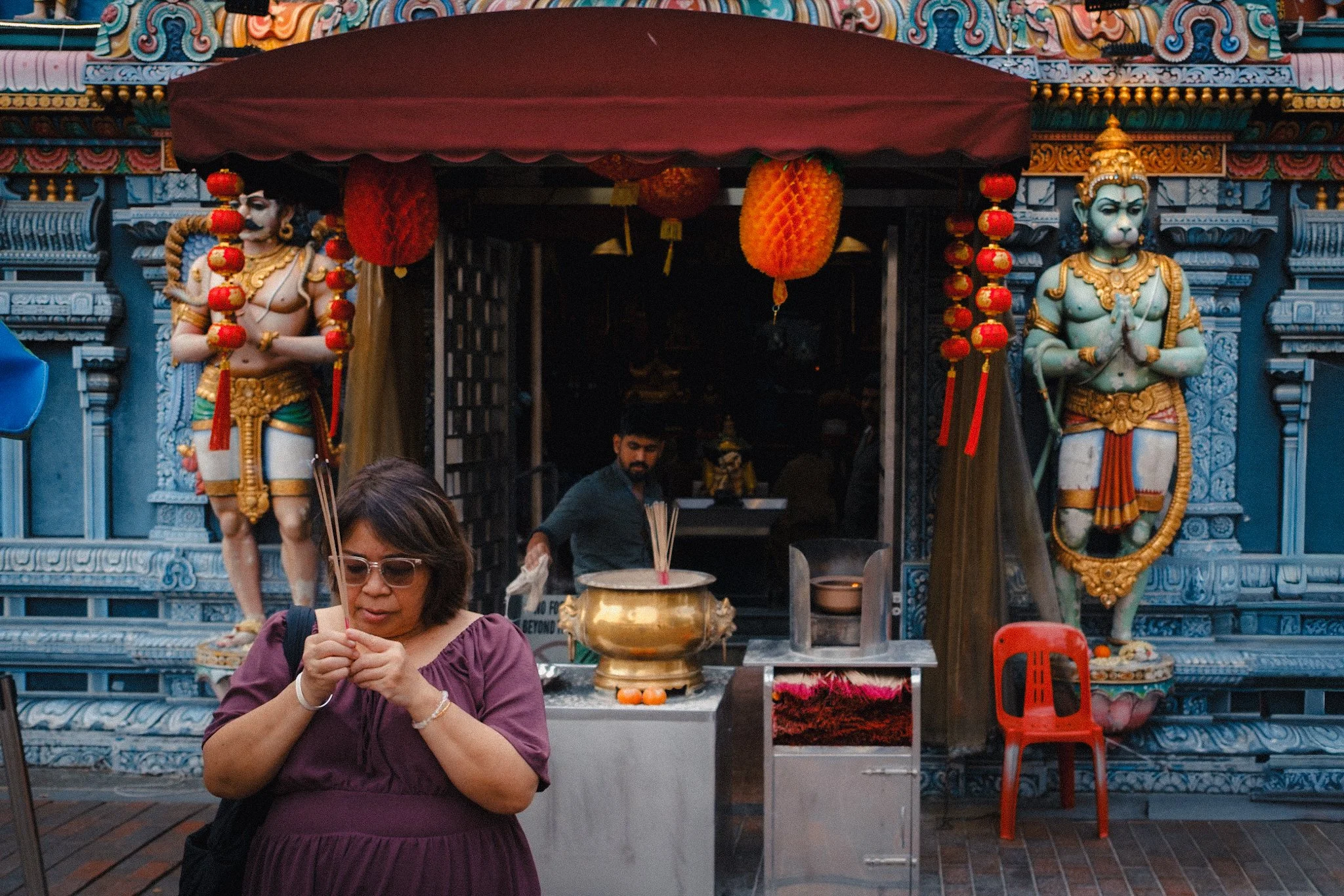 A woman in a purple dress holding incense sticks stands in front of a Hindu temple entrance decorated with statues of deities, lanterns, and flowers, with a man behind her preparing offerings.