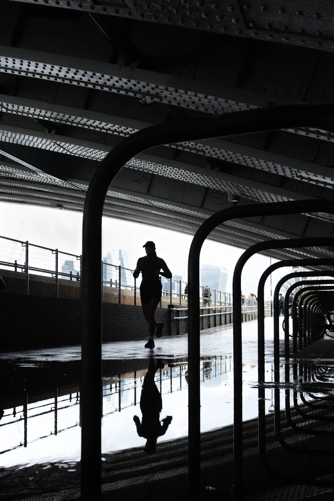 A person running under a bridge, with their reflection visible in a puddle on the ground. The background shows a city skyline with tall buildings.