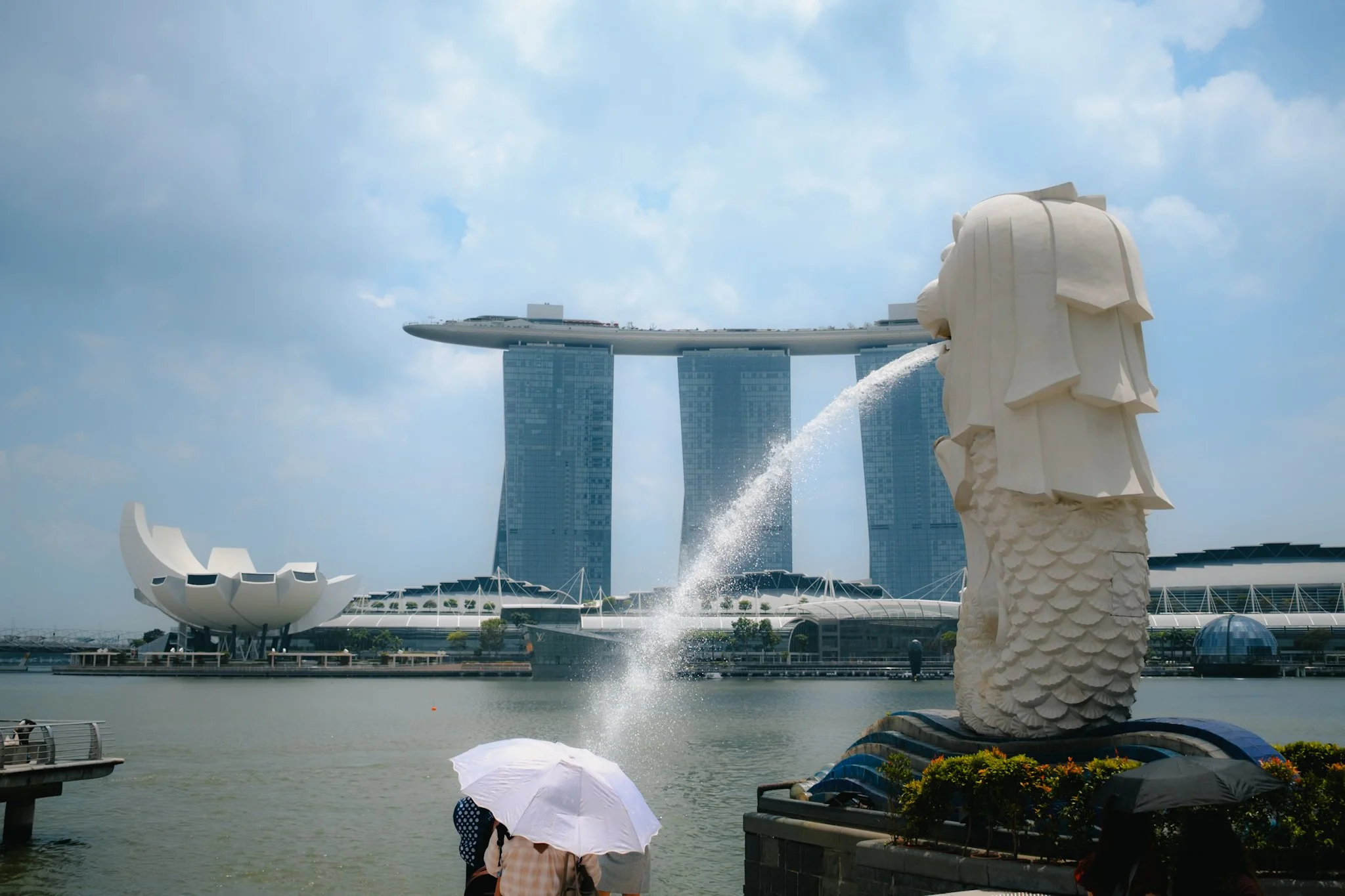 View of Marina Bay Sands hotel, Merlion statue, and ArtScience Museum in Singapore with people holding umbrellas in the foreground.