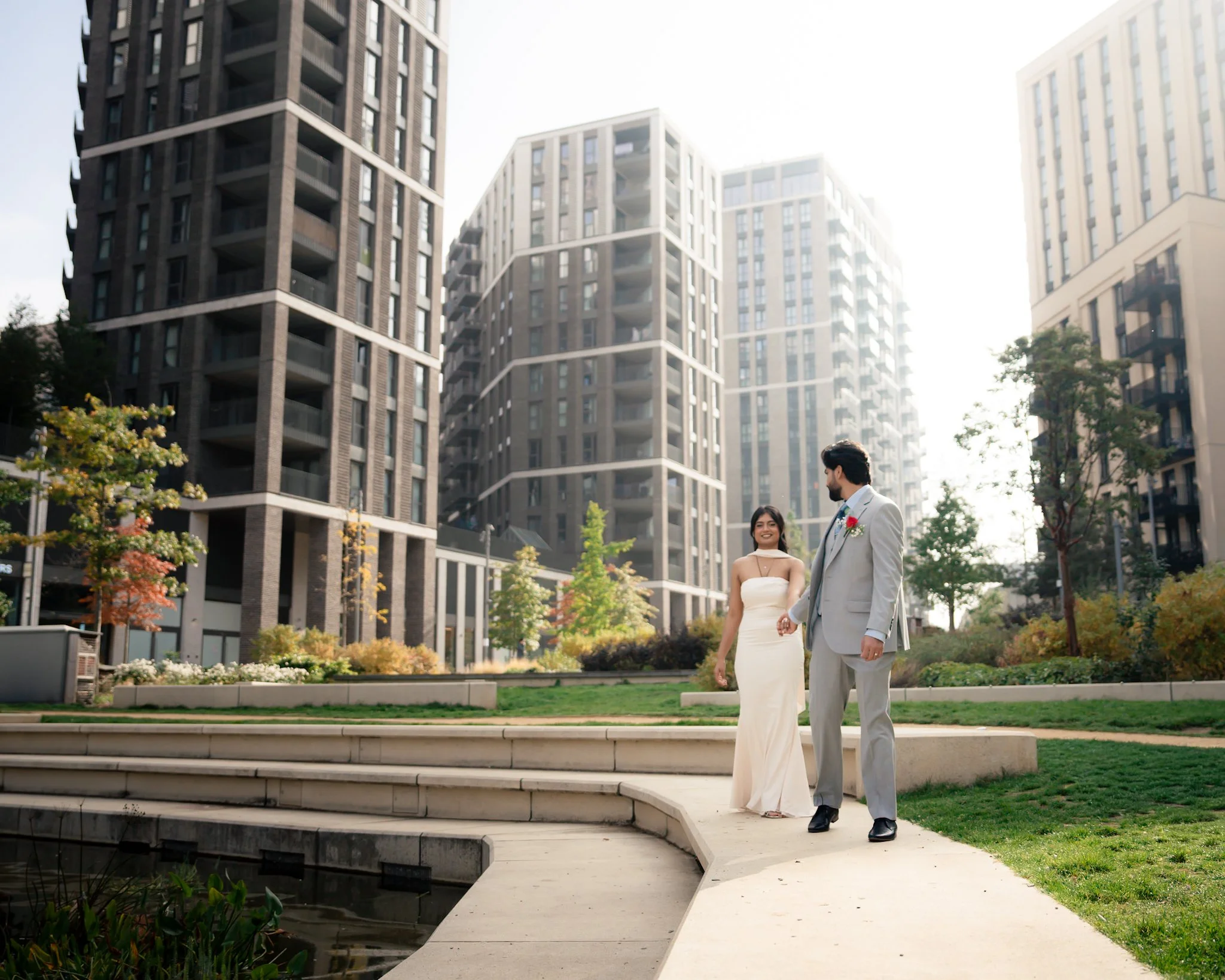 A couple dressed in wedding attire, standing outdoors on steps near a pond, with modern high-rise buildings in the background on a bright, sunny day.