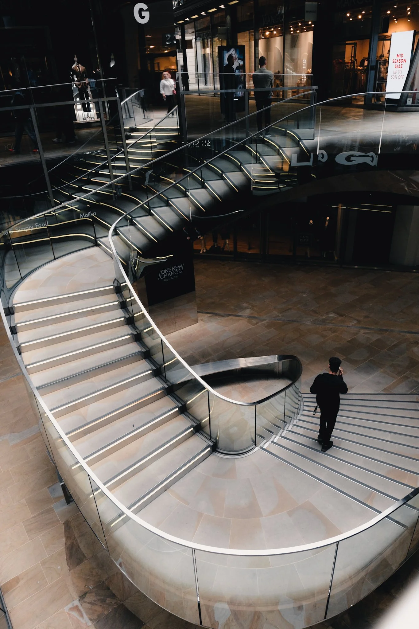 Interior view of a modern shopping mall featuring a curved escalator with glass sides and illuminated steps, a staircase with matching design, and several people walking or standing. Stores and digital advertisements are visible in the background.