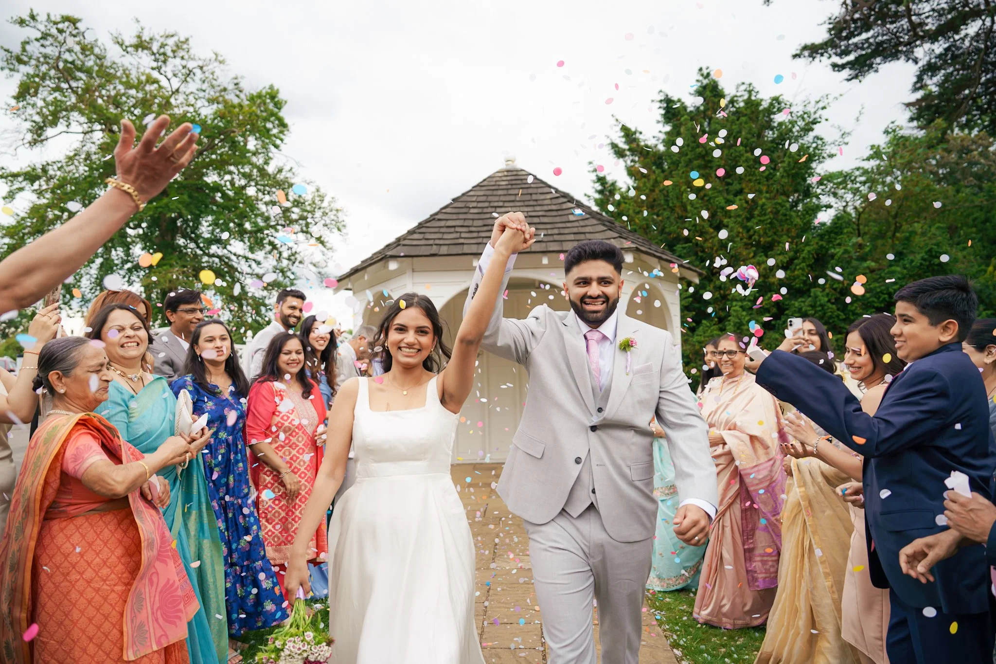 A diverse group of people celebrating a wedding outdoors. The bride and groom are smiling and walking together, with the groom raising his fist in celebration. Colorful confetti is falling around them, and guests are clapping and taking photos. There