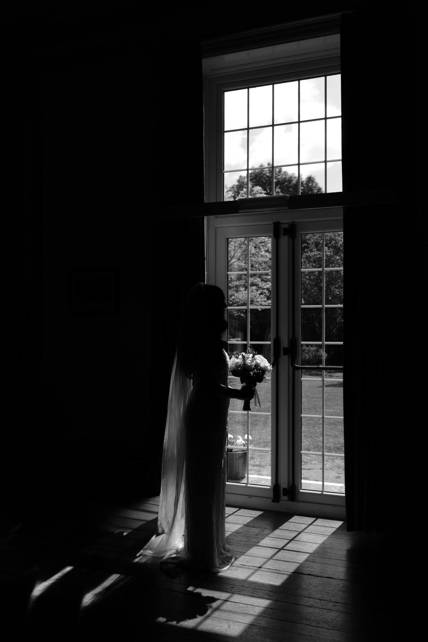 A woman in a wedding dress holding a bouquet stands near a large window, with sunlight casting shadows on the wooden floor, creating a silhouette effect.