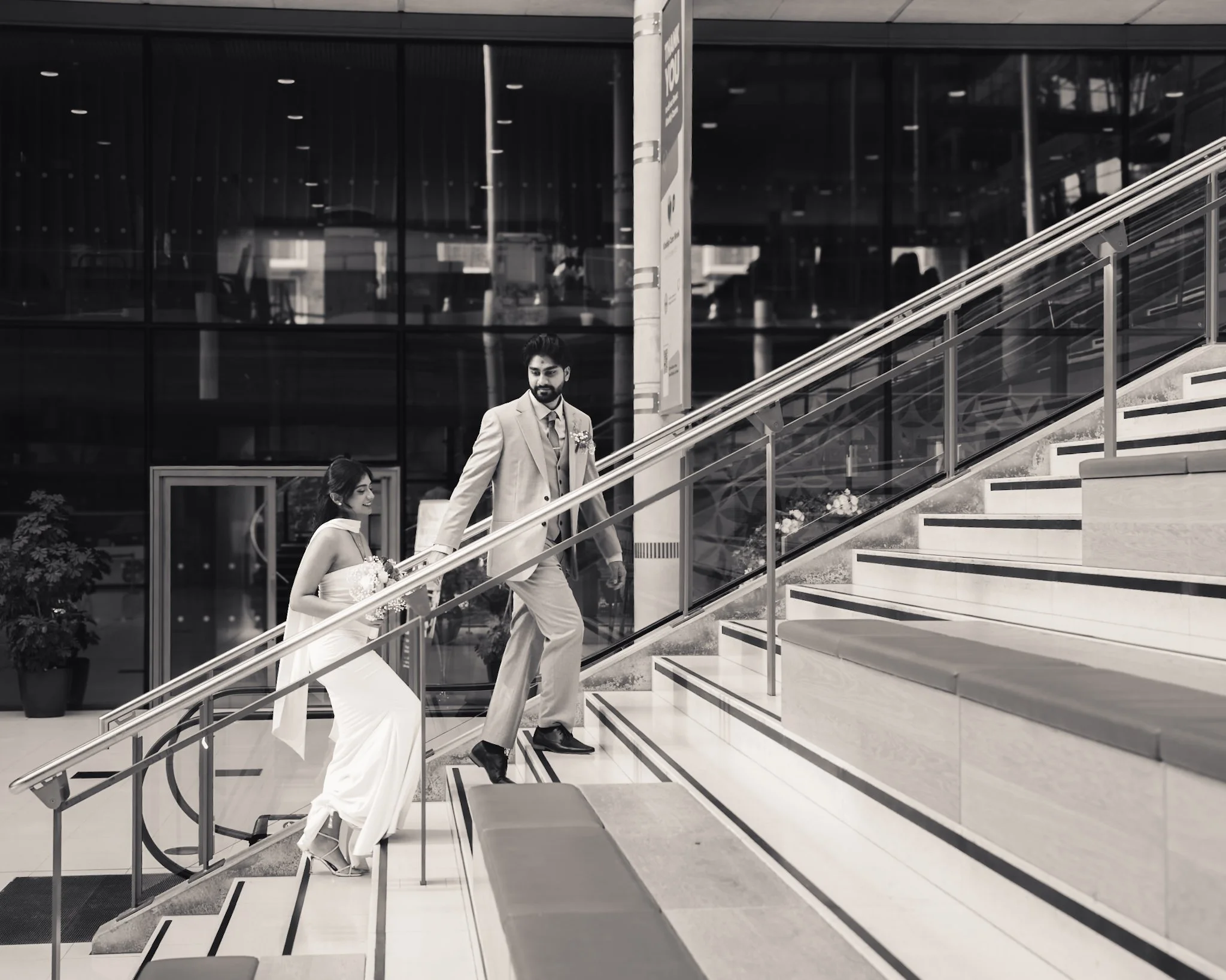 A man and woman in wedding attire ascending a staircase inside a modern building with glass walls and urban reflections in black and white.
