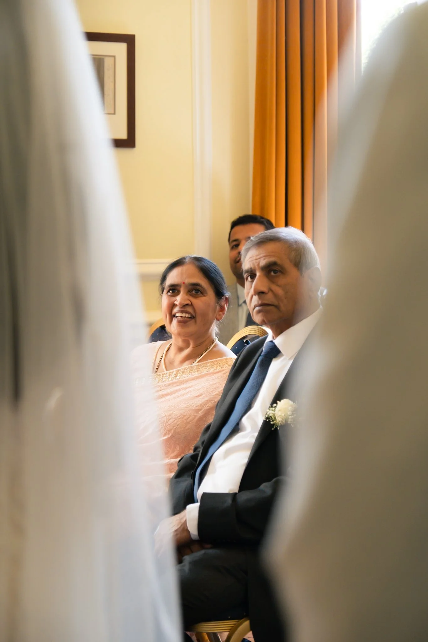 People seated in a formal setting, seen through a narrow opening, with a woman wearing traditional Indian attire and a man in a suit with a white flower boutonniere.