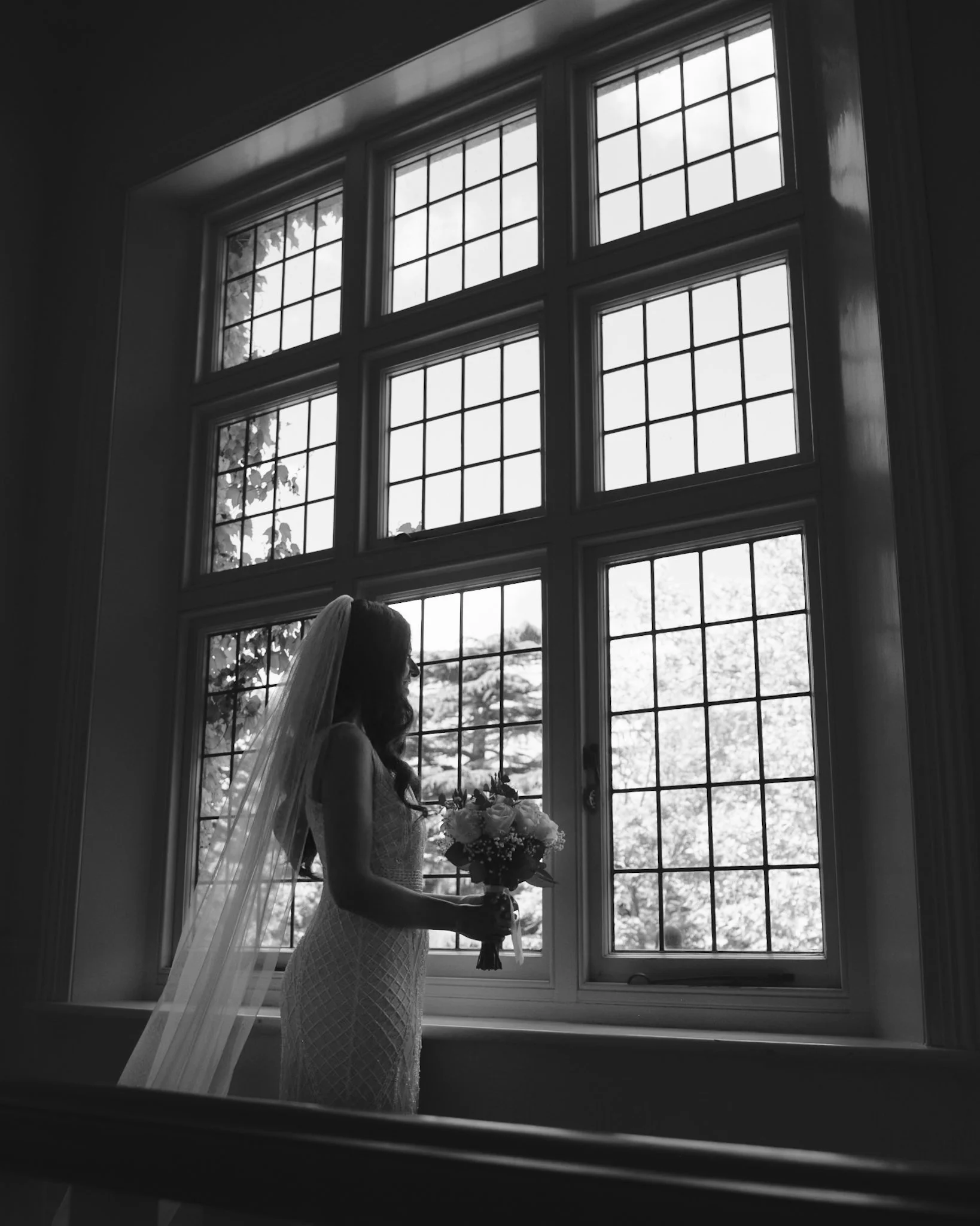 A silhouette of a bride holding a bouquet of flowers, standing in front of a large window with multiple panes, with light streaming through the window and a tree visible outside.