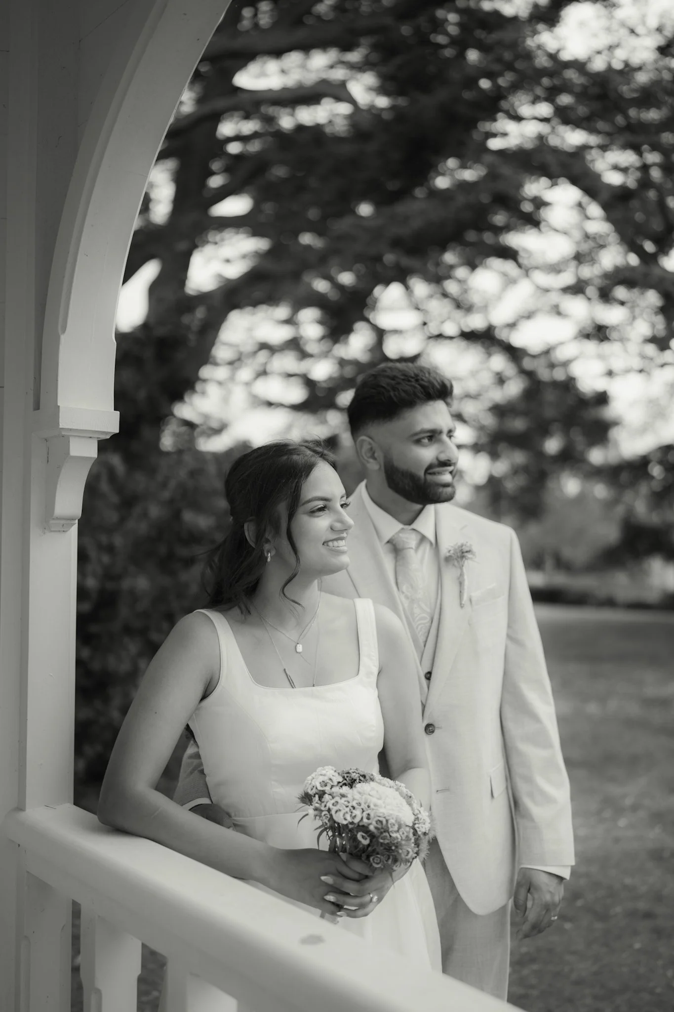 A black and white photo of a bride and groom standing outdoors. The bride is holding a bouquet of flowers and wearing a sleeveless wedding dress. The groom is dressed in a light-colored suit and tie. They are visible through a white decorative arch, 