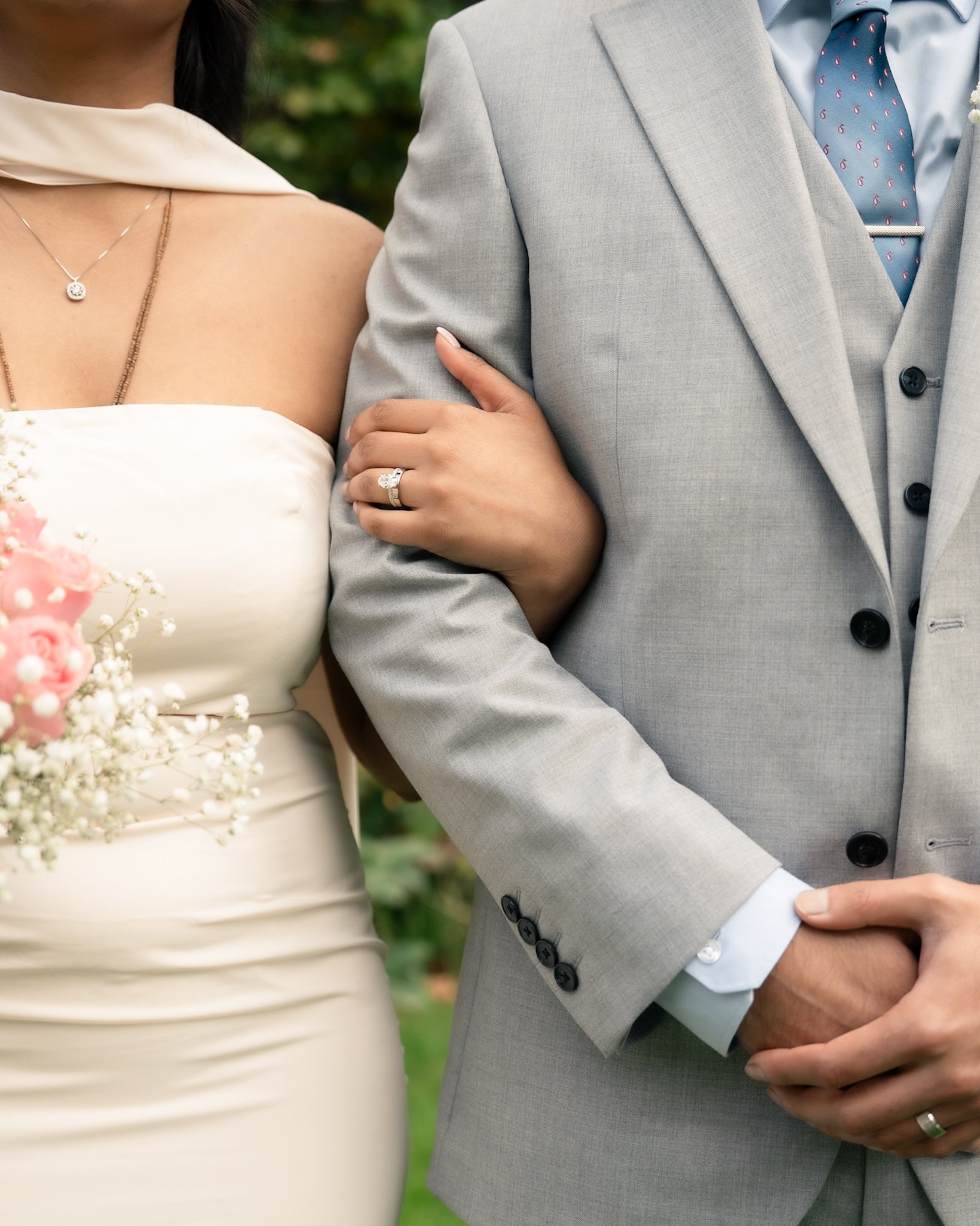 Close-up of a couple dressed in wedding attire, with the woman wearing a white dress and jewelry, and the man in a light gray suit with a blue tie, standing closely outdoors.