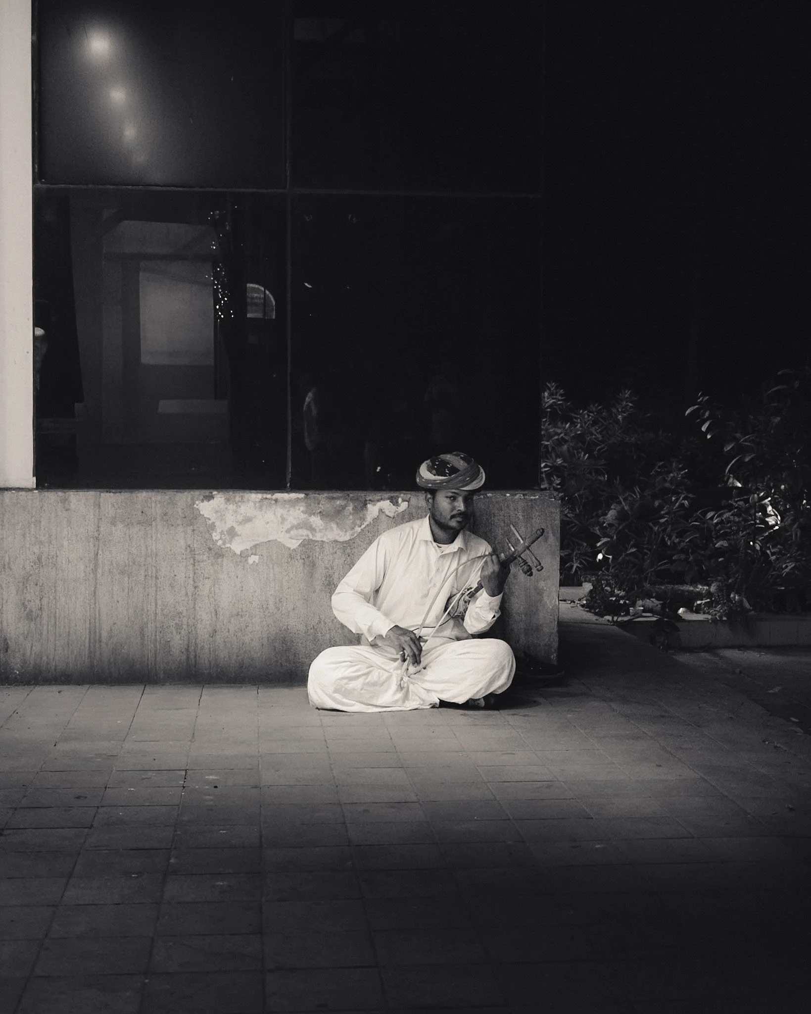 A man in traditional white attire, sitting on the ground and playing a violin, outside at night. Behind him, there is a wall, some plants, and a window with reflections.