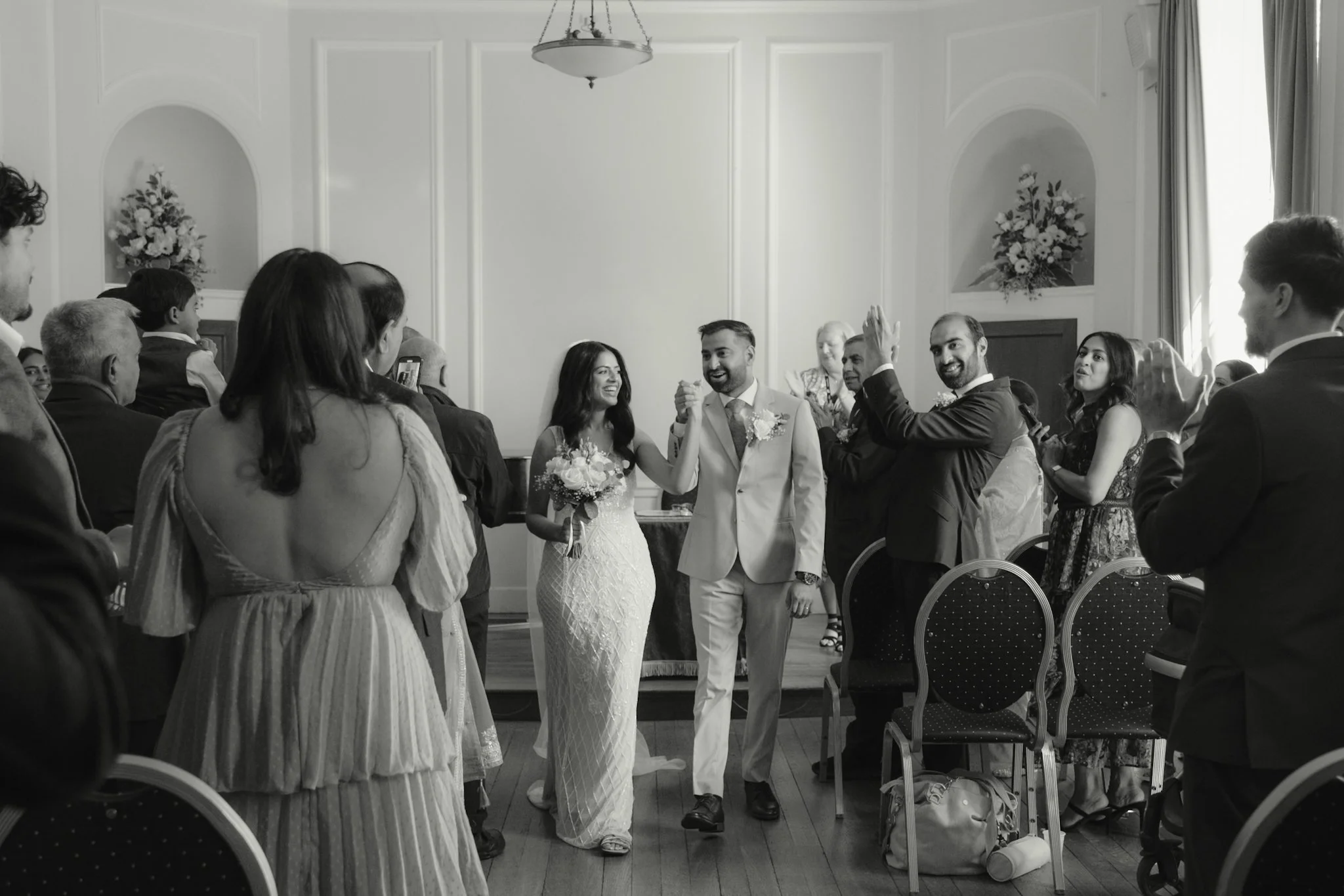 A wedding ceremony with the bride and groom holding hands and smiling, surrounded by guests. The bride is wearing a white wedding dress and holding a bouquet, while the groom is dressed in a light-colored suit. Guests are sitting on chairs and watchi