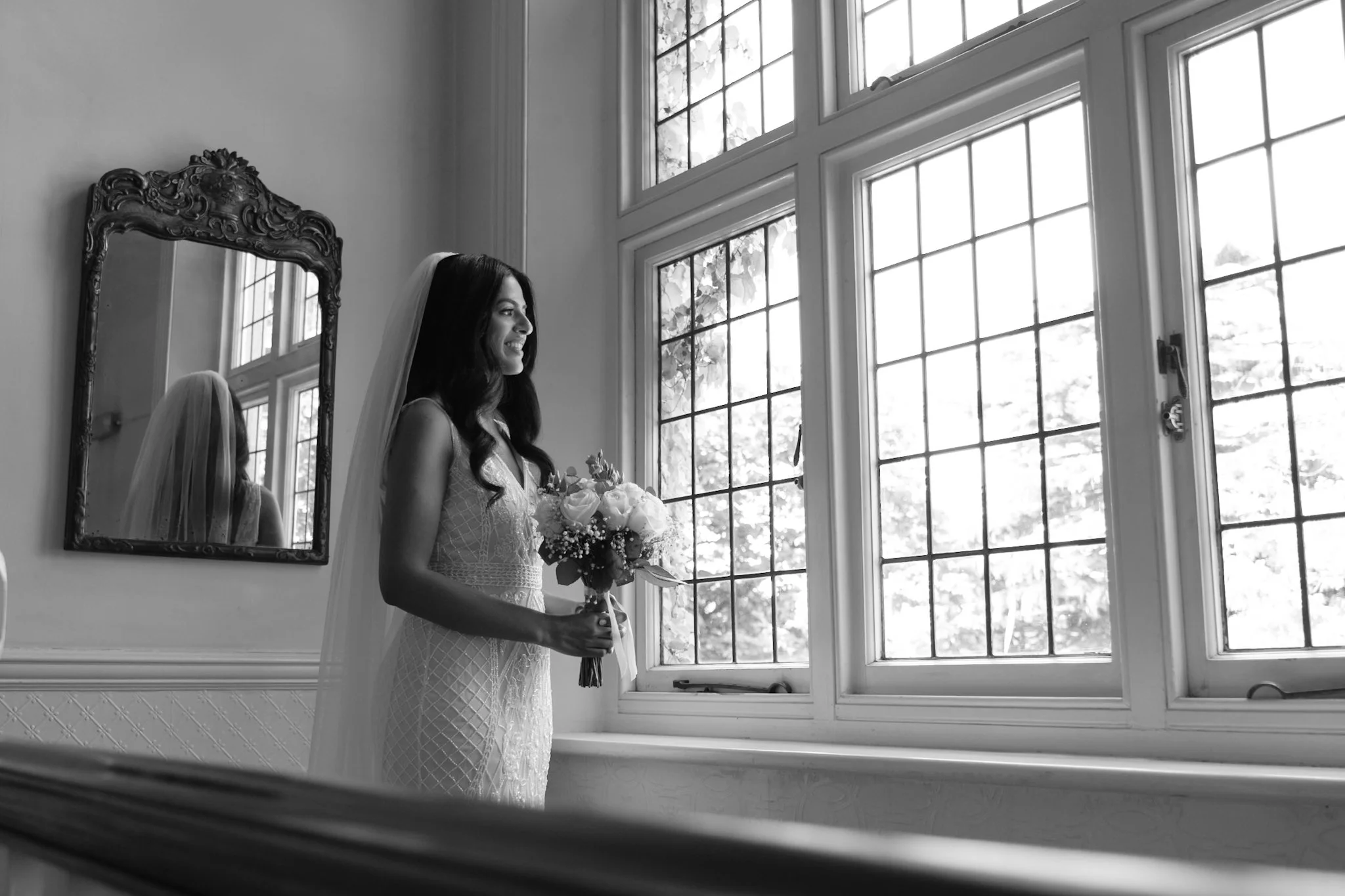 A bride with dark hair in a lace wedding dress and veil holding a bouquet of flowers, standing near a large window with grid panes, reflected in an ornate mirror on the wall.