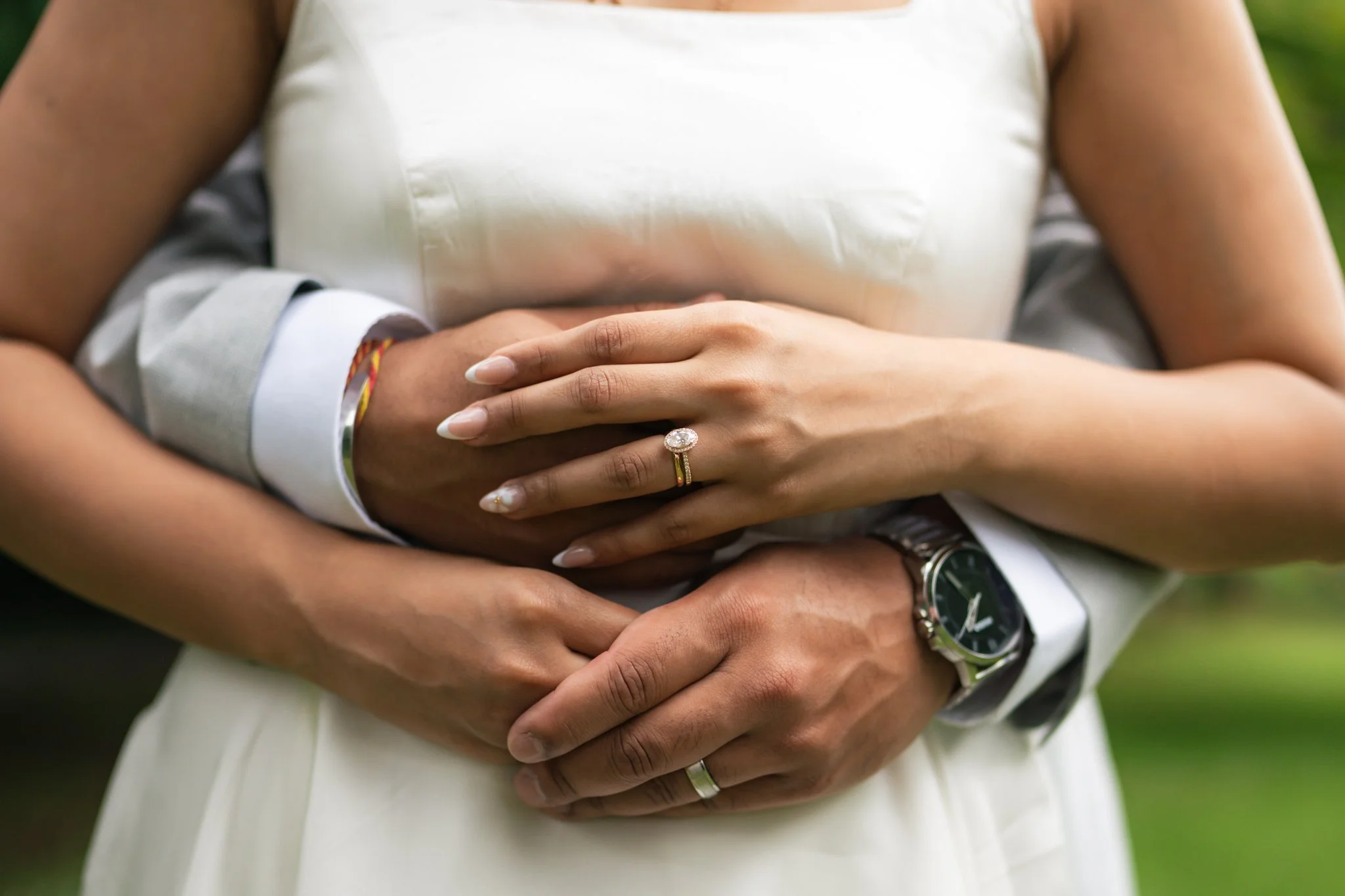 A close-up of two people embracing, with their arms crossed over each other, one wearing a wedding ring and the other a watch, in an outdoor setting.