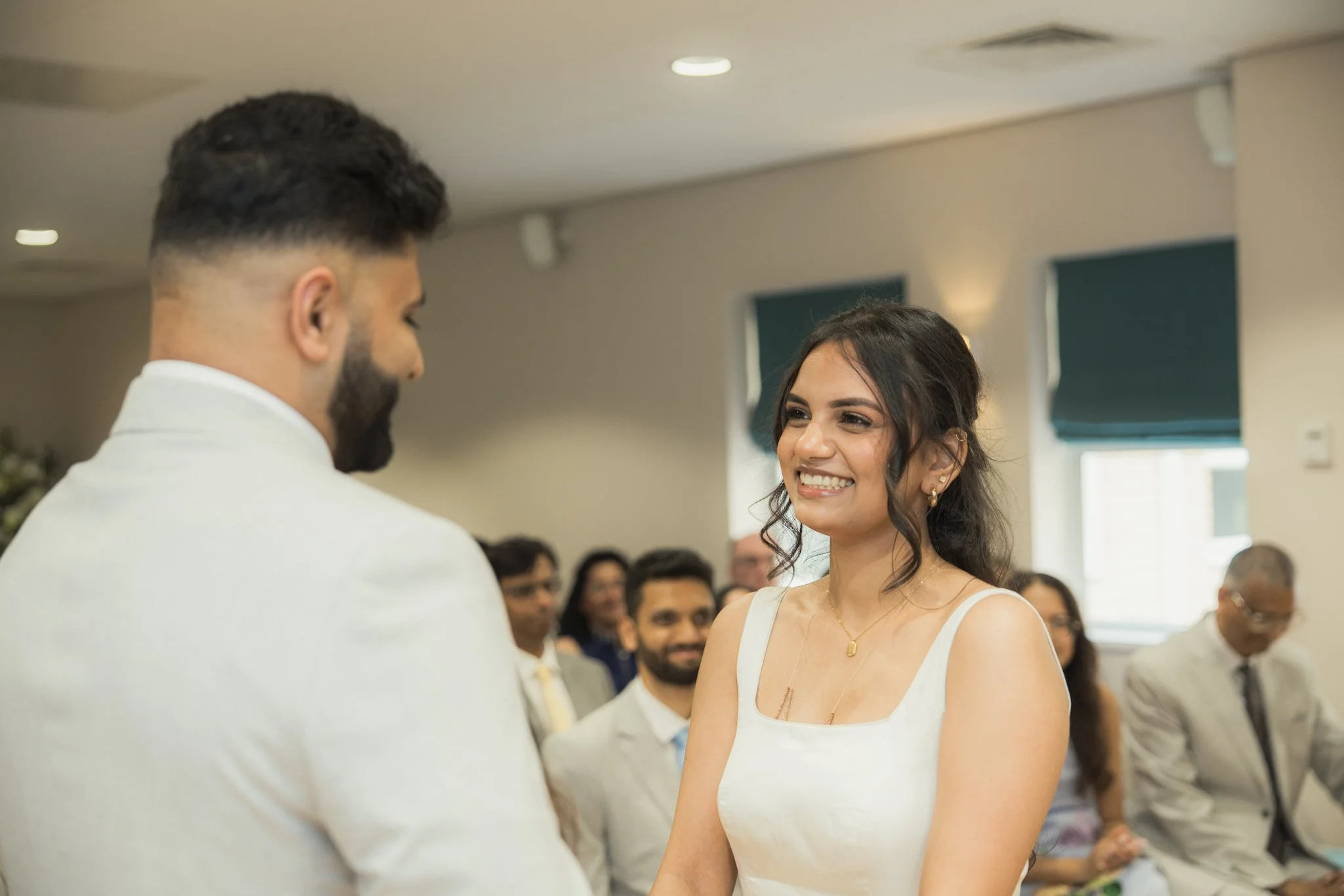 A woman in a white dress is smiling at a man in a white blazer during a wedding ceremony. The background shows a seated crowd of wedding guests.