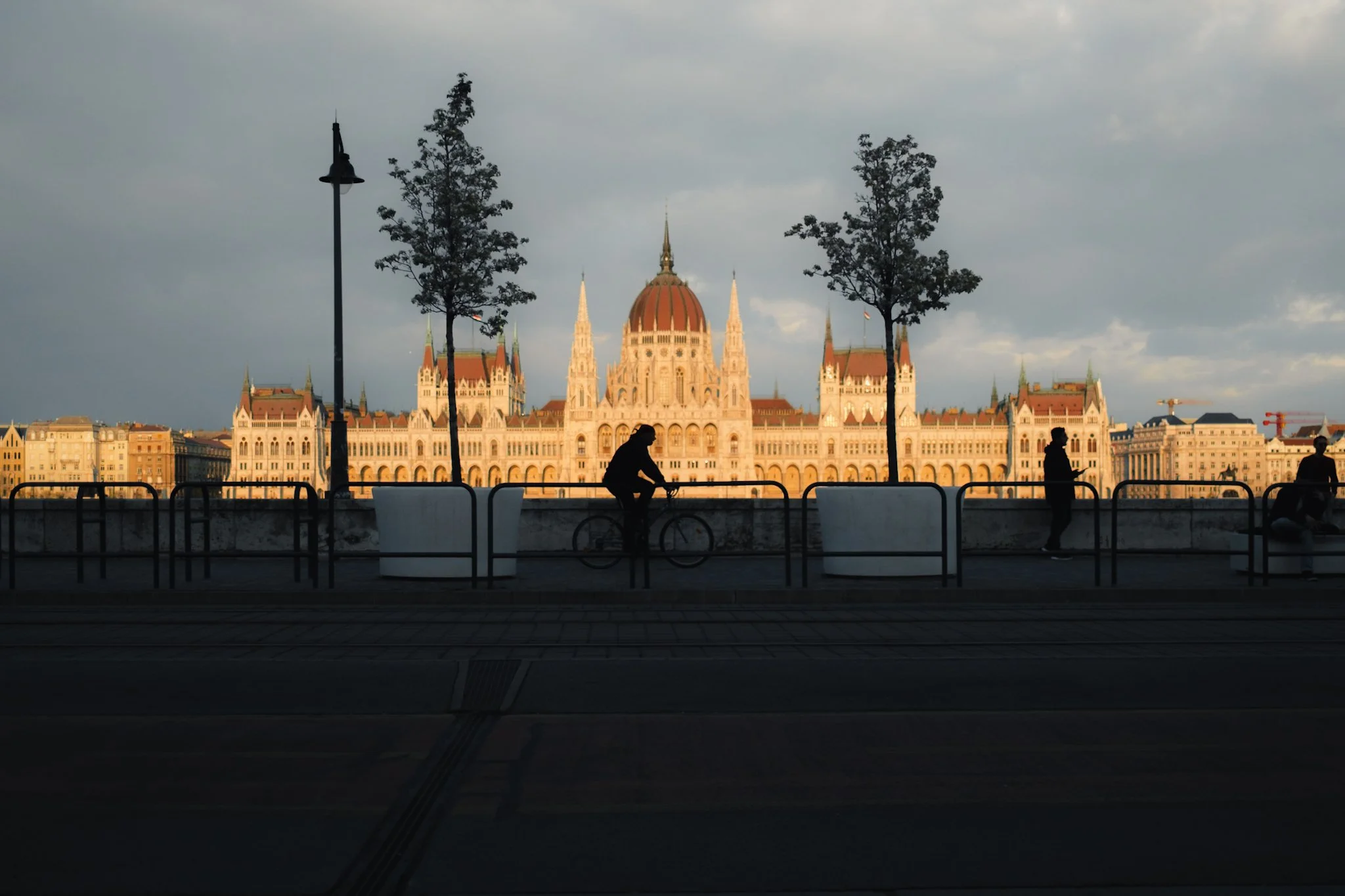 View of the Hungarian Parliament Building in Budapest with a cloudy sky, silhouetted people, and trees in the foreground.