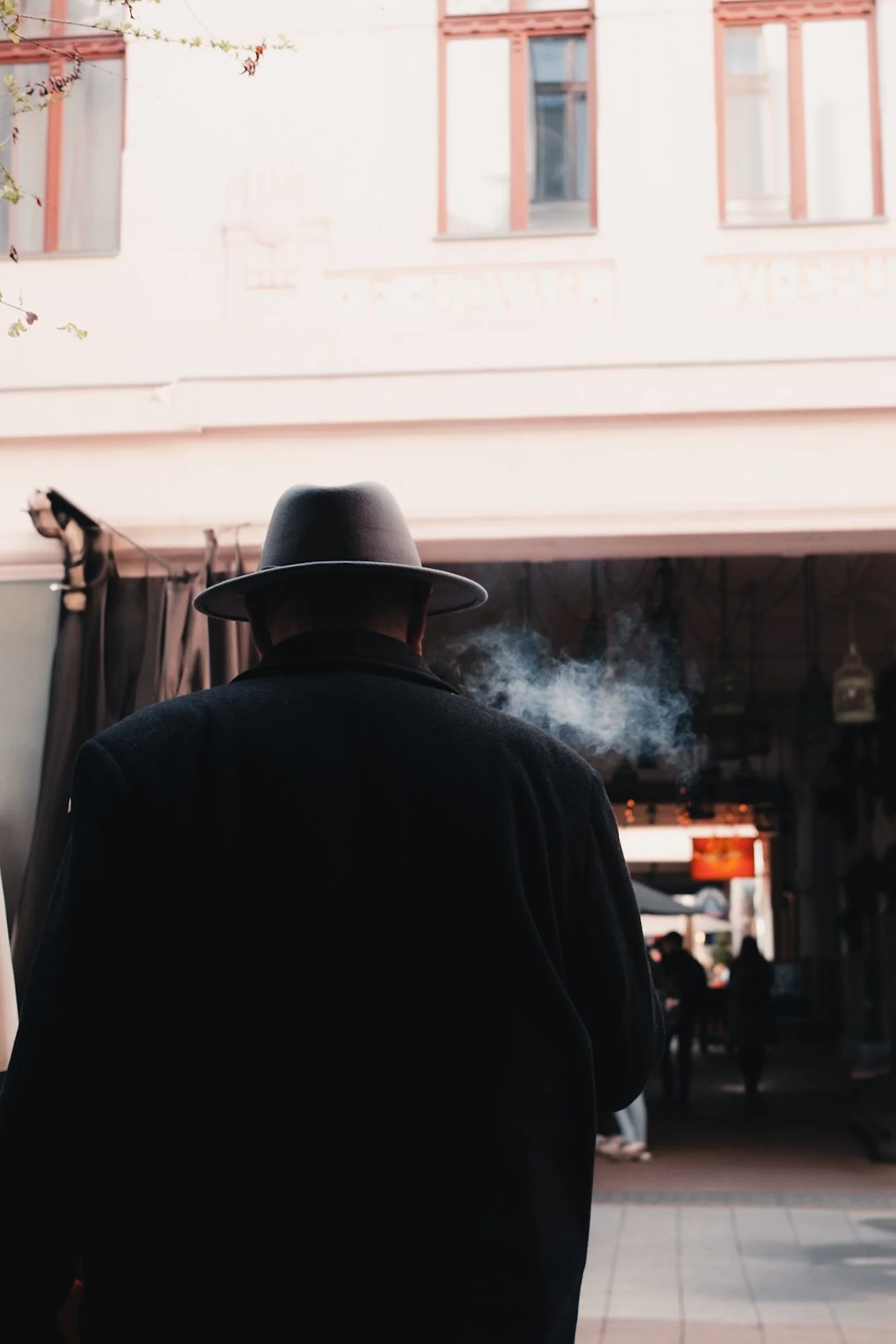 A man wearing a black coat and a fedora hat, smoking, facing away from the camera, in an indoor space with a view of a building with windows and some people in the background.