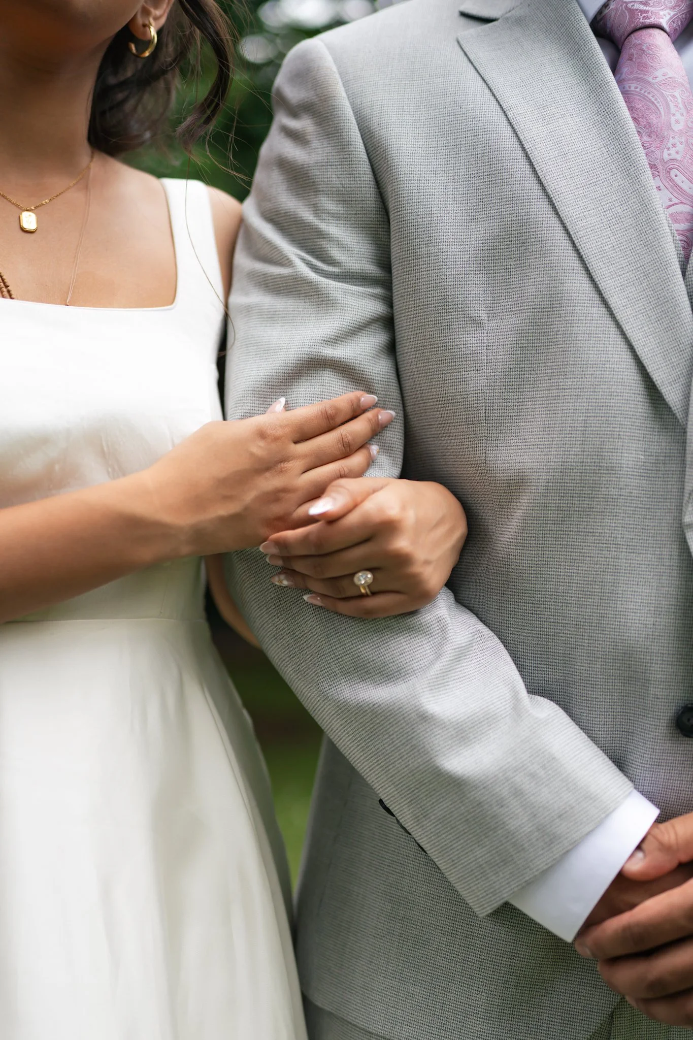 Close-up of a woman and man holding hands, dressed in formal wedding attire. The woman wears a white dress and gold jewelry, while the man wears a gray suit with a pink patterned tie.