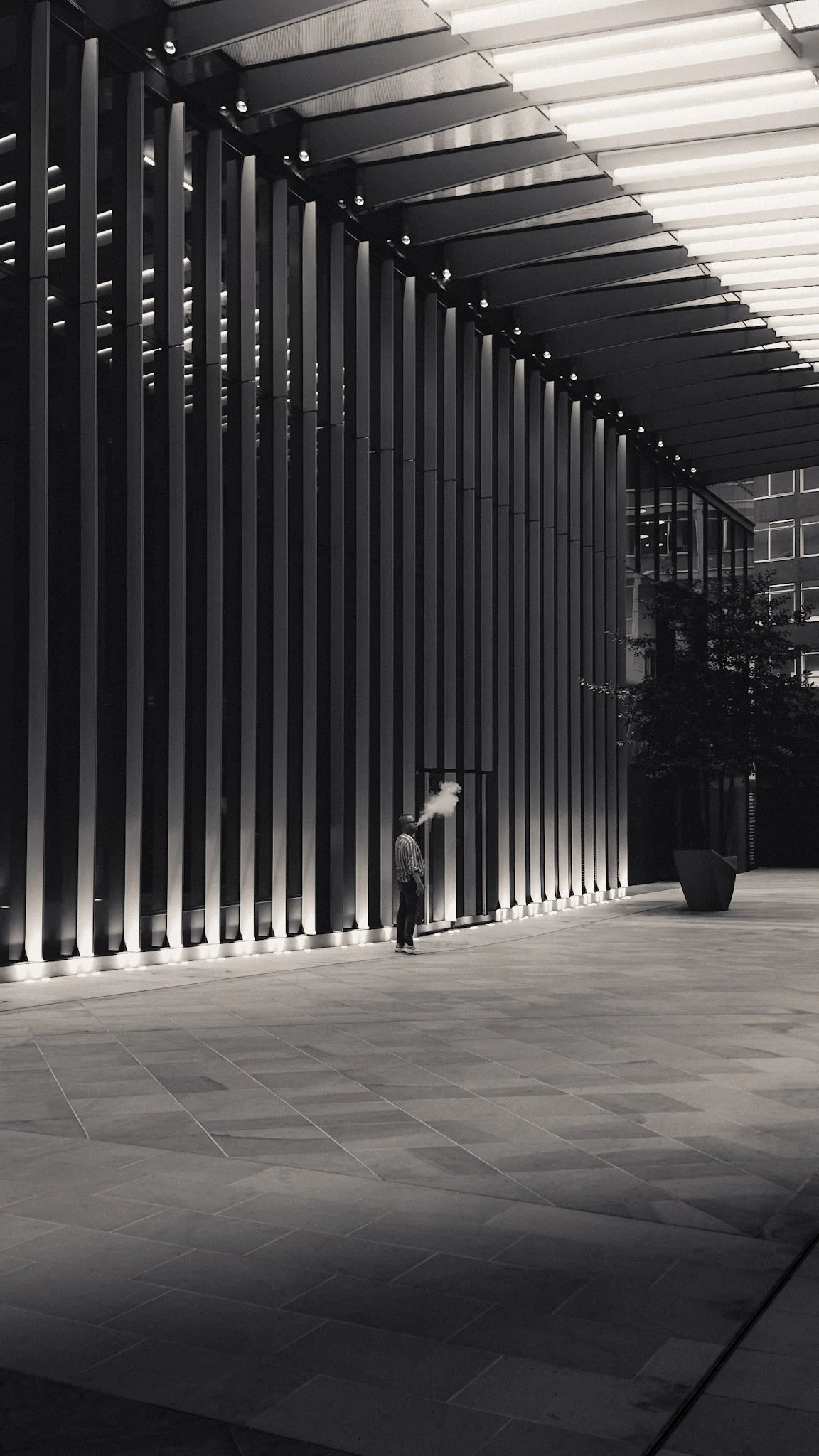 A person standing outside a modern building with vertical metal slats on the facade, smoking and exhaling vapor in an urban setting at night.