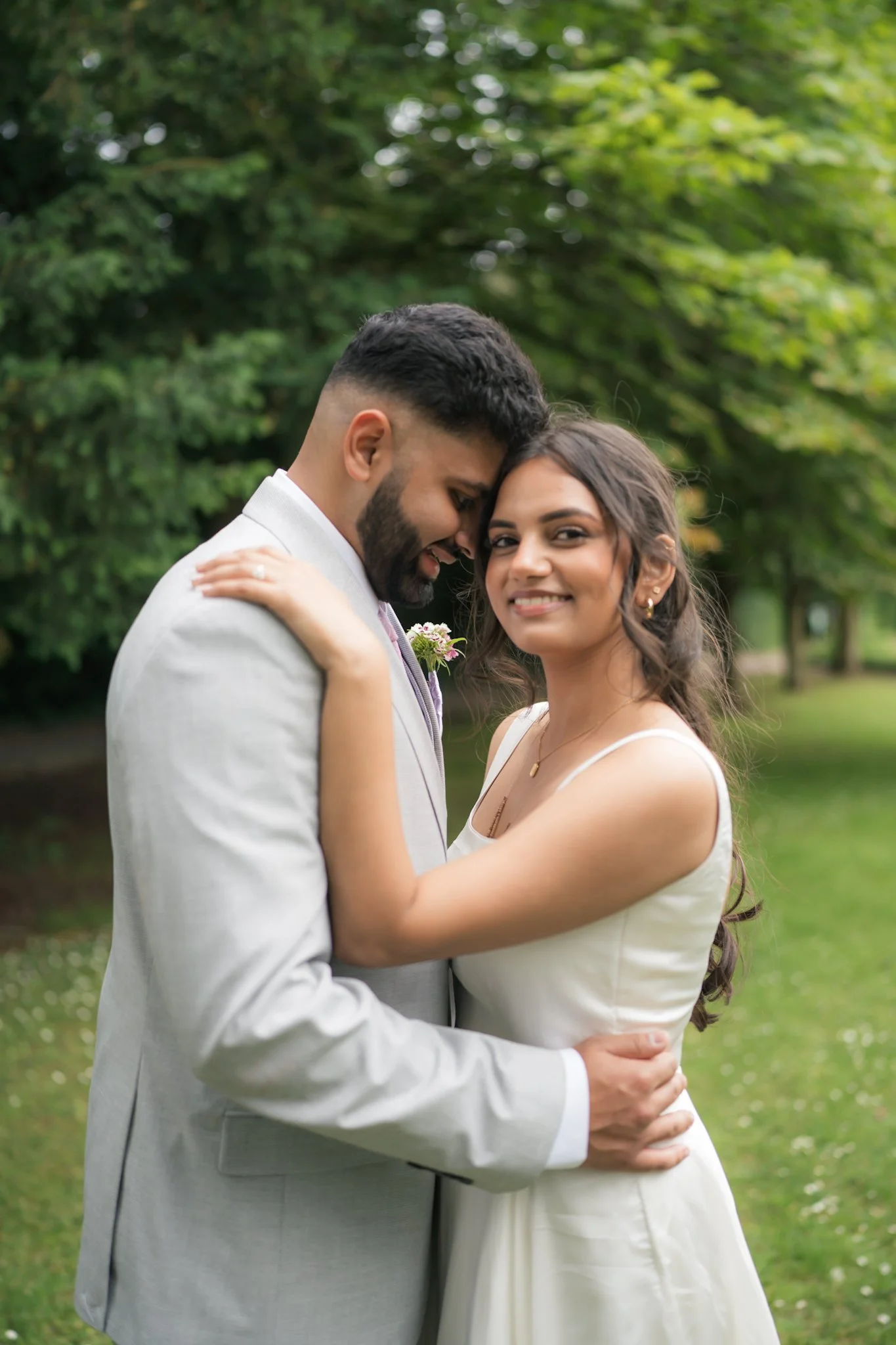 A bride and groom embrace outdoors with green trees in the background. The groom is wearing a light gray suit and the bride is dressed in a white wedding gown. They are holding each other close, smiling softly.