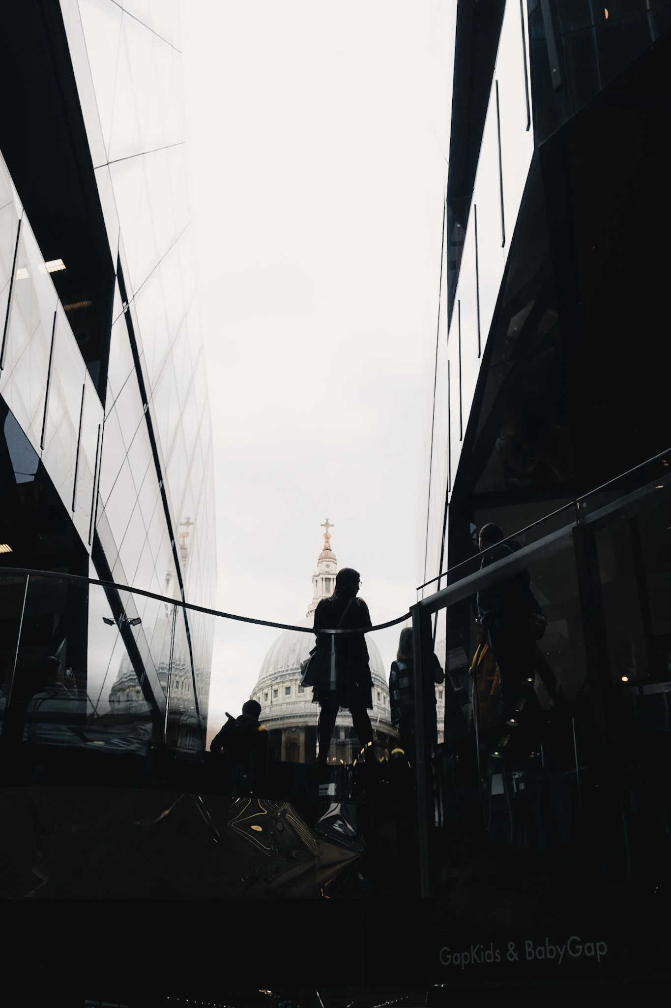 People walking inside a modern shopping mall with glass walls, with a view of a historic dome and clock tower outside through the opening.