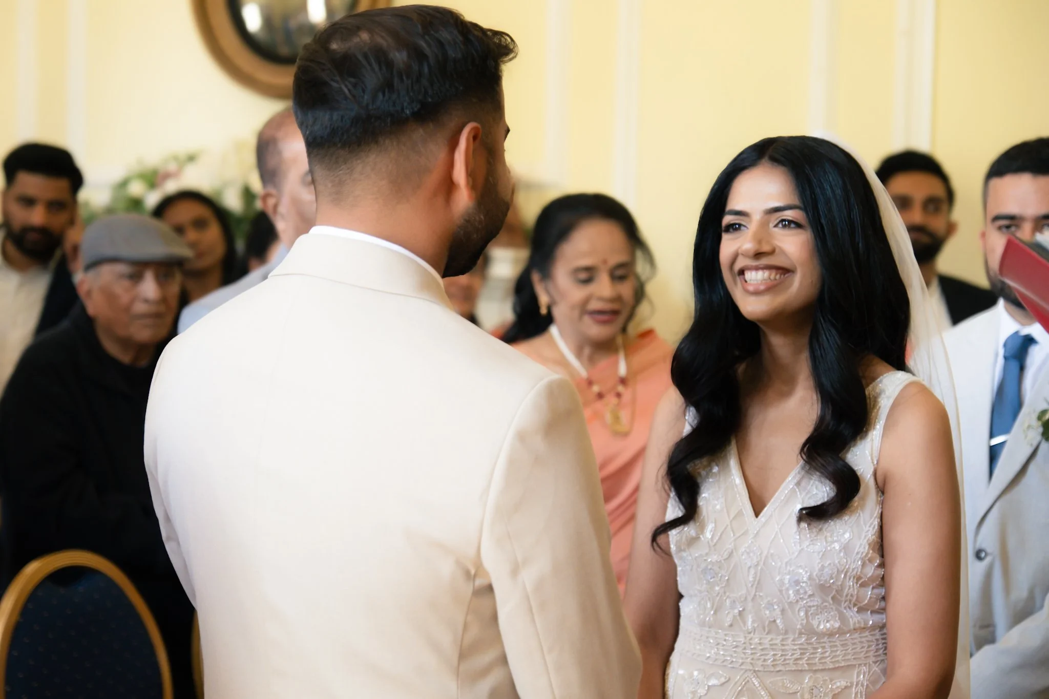A woman in a white dress with black hair smiling at a man in a cream-colored suit during a wedding ceremony with guests in the background.