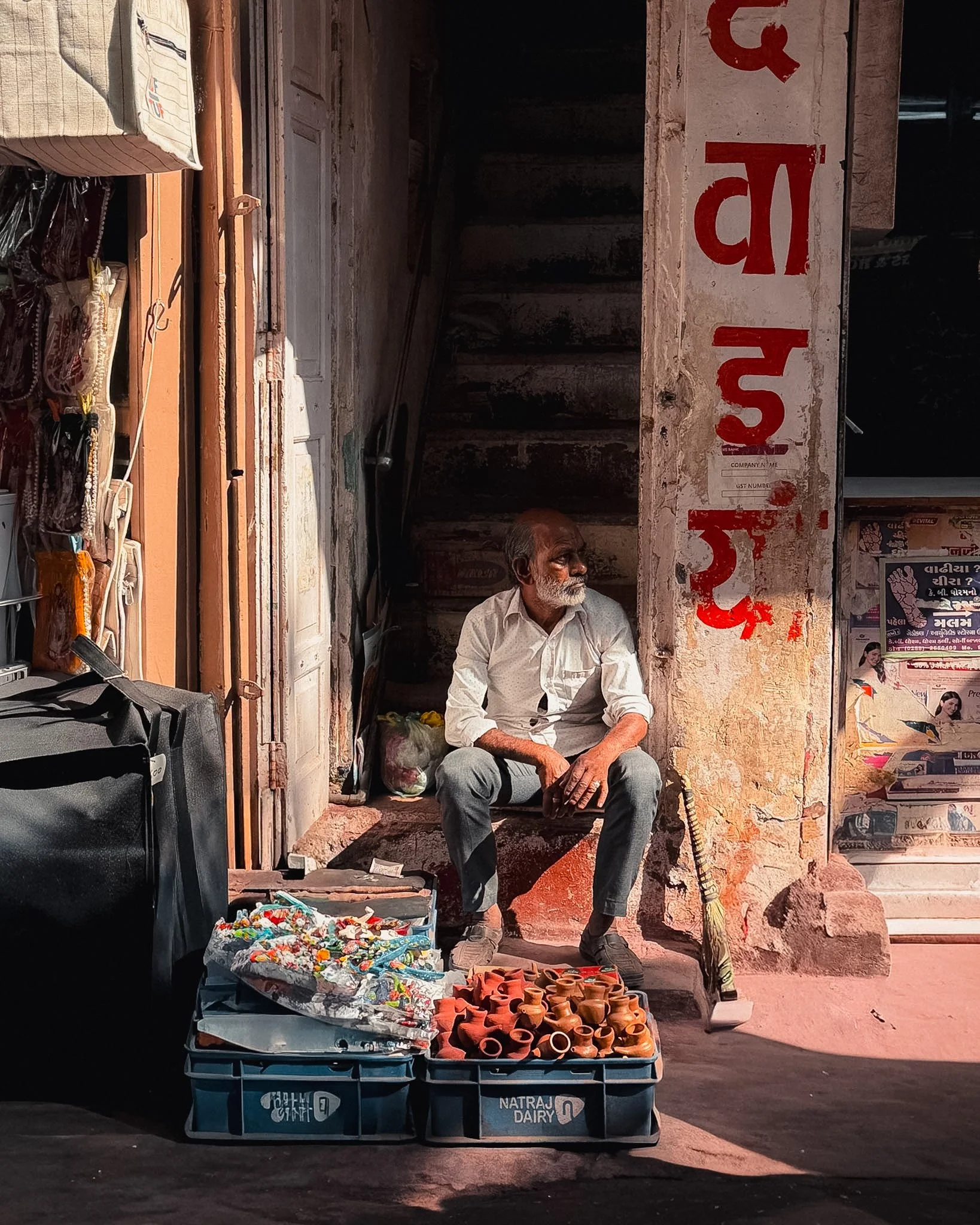 An elderly man sitting on a chair on the street with various small clay pots on a cart in front of him, a woman behind him, and a sign with red text on the wall behind him.