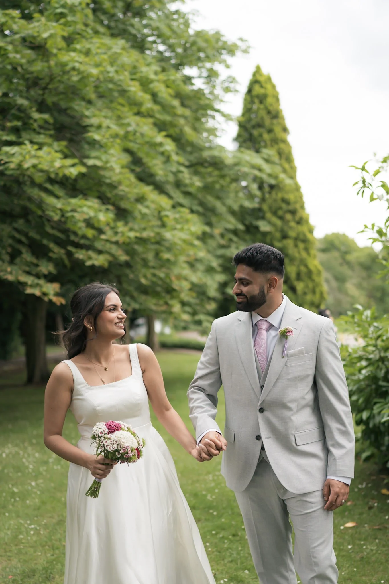 A bride and groom holding hands and walking outdoors in a park, surrounded by green trees, with the bride holding a bouquet of flowers and smiling at the groom.