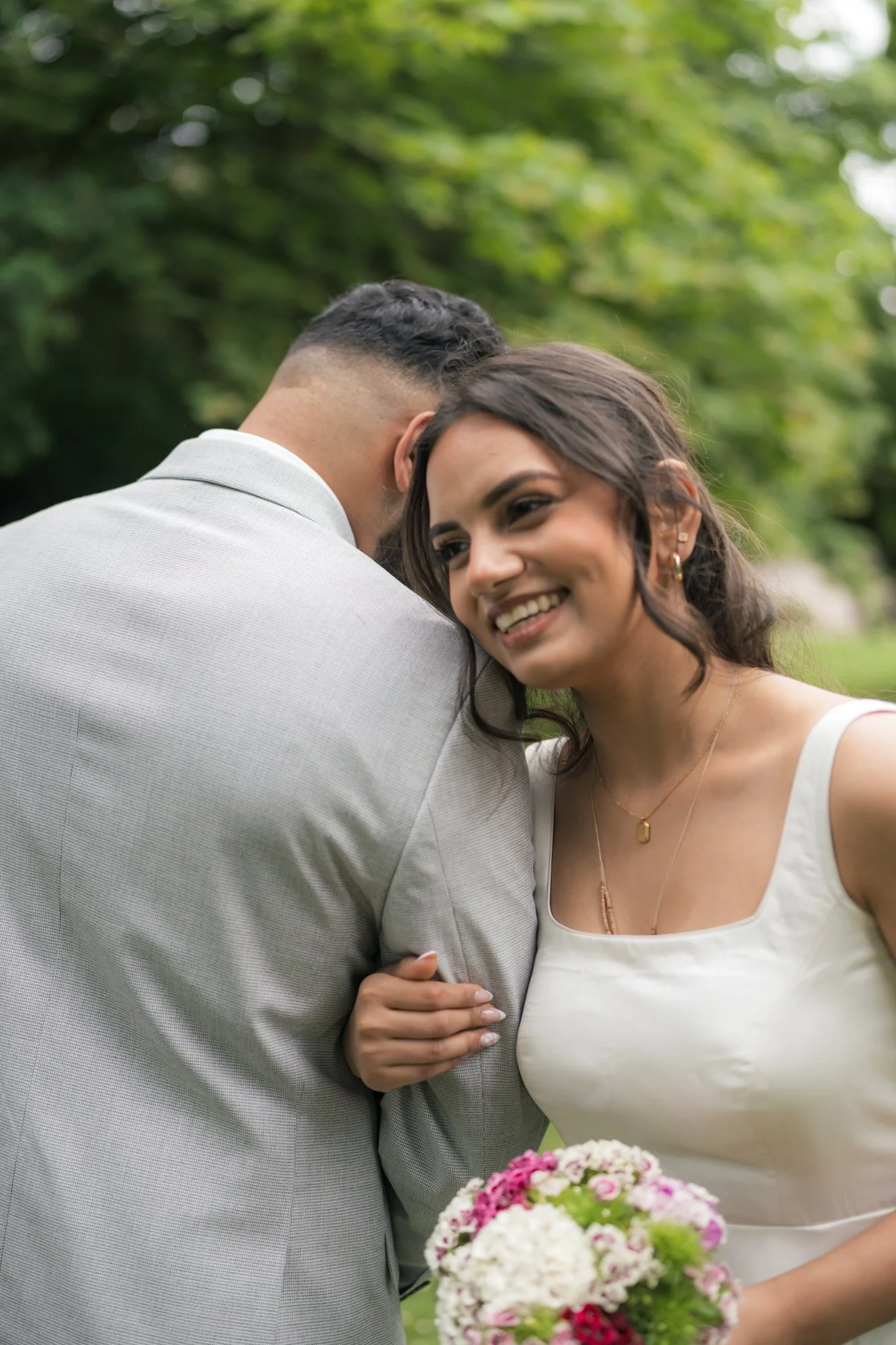 A smiling woman holding a bouquet of mixed pink and white flowers, standing outdoors with a man, both dressed casually, amidst greenery.