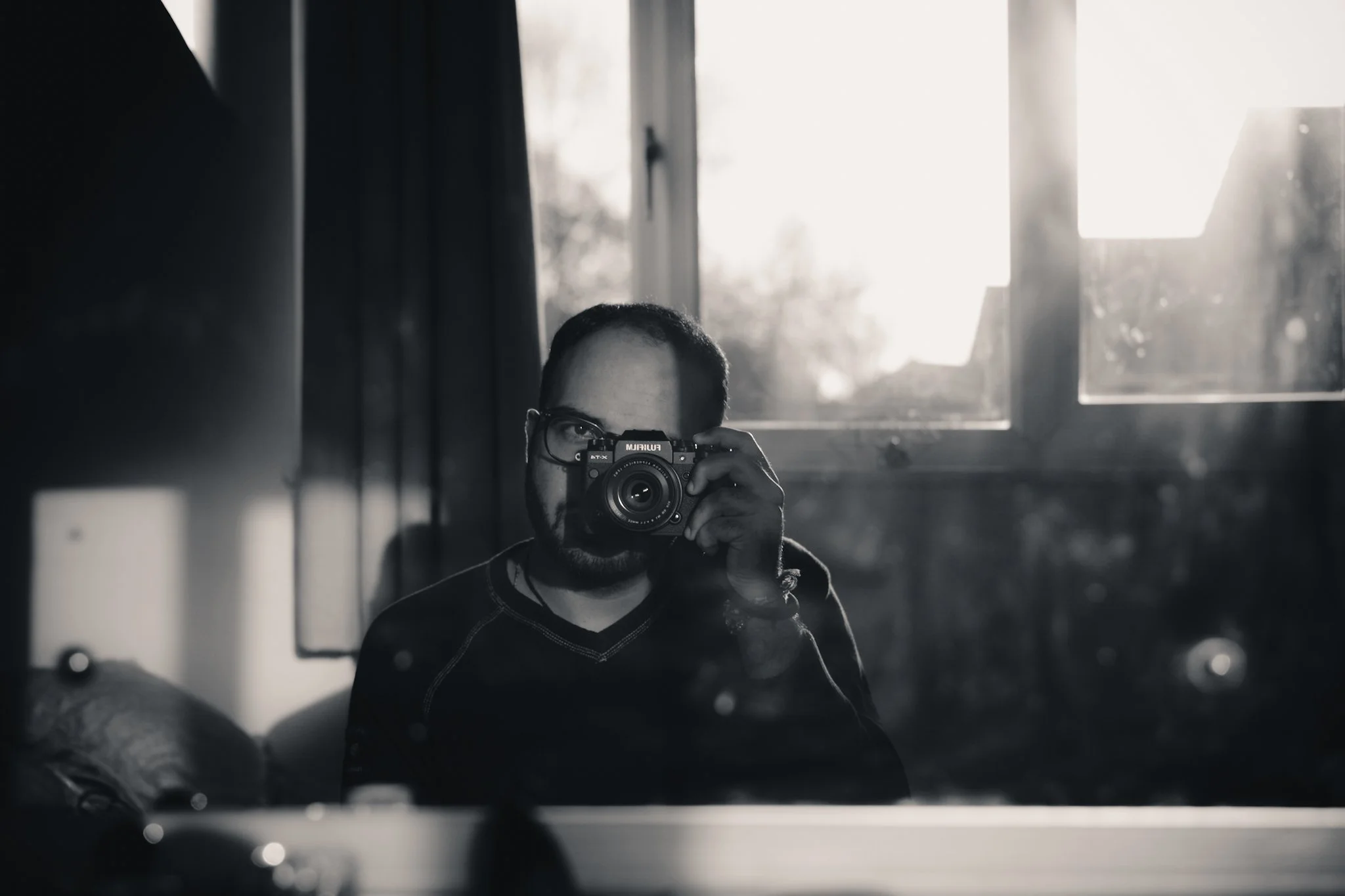 A man with glasses taking a photo of himself with a camera in a room with sunlight coming through a window.