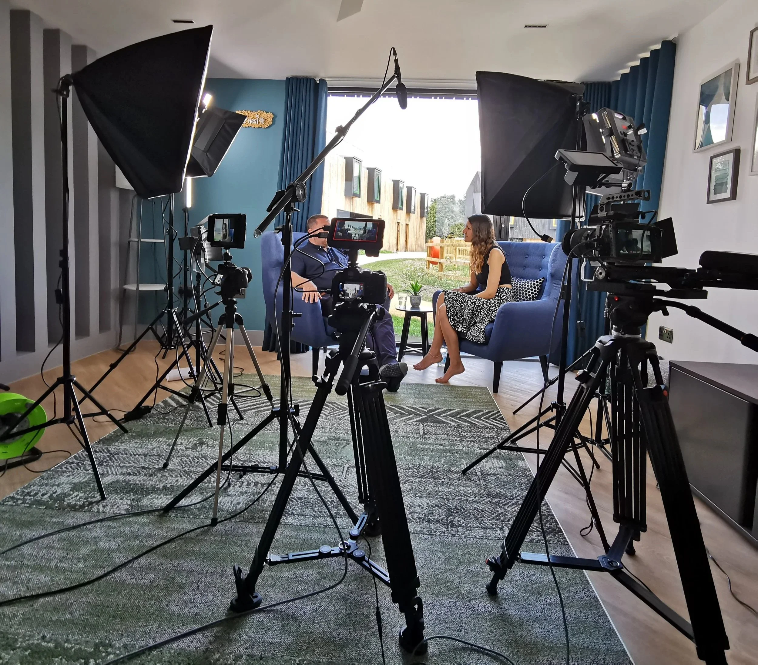 A recording setup with cameras and lighting equipment directed at two people sitting on blue chairs, engaged in conversation in a well-lit room with large windows and framed artwork on the wall.