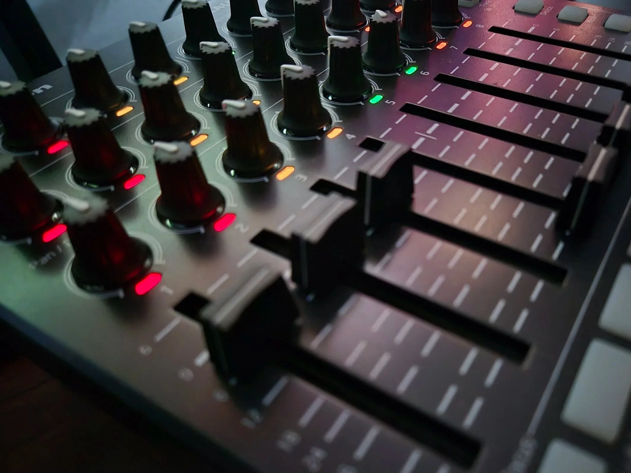 Close-up of a professional audio mixer with multiple knobs, sliders, and indicator lights illuminated in red, orange, and green.