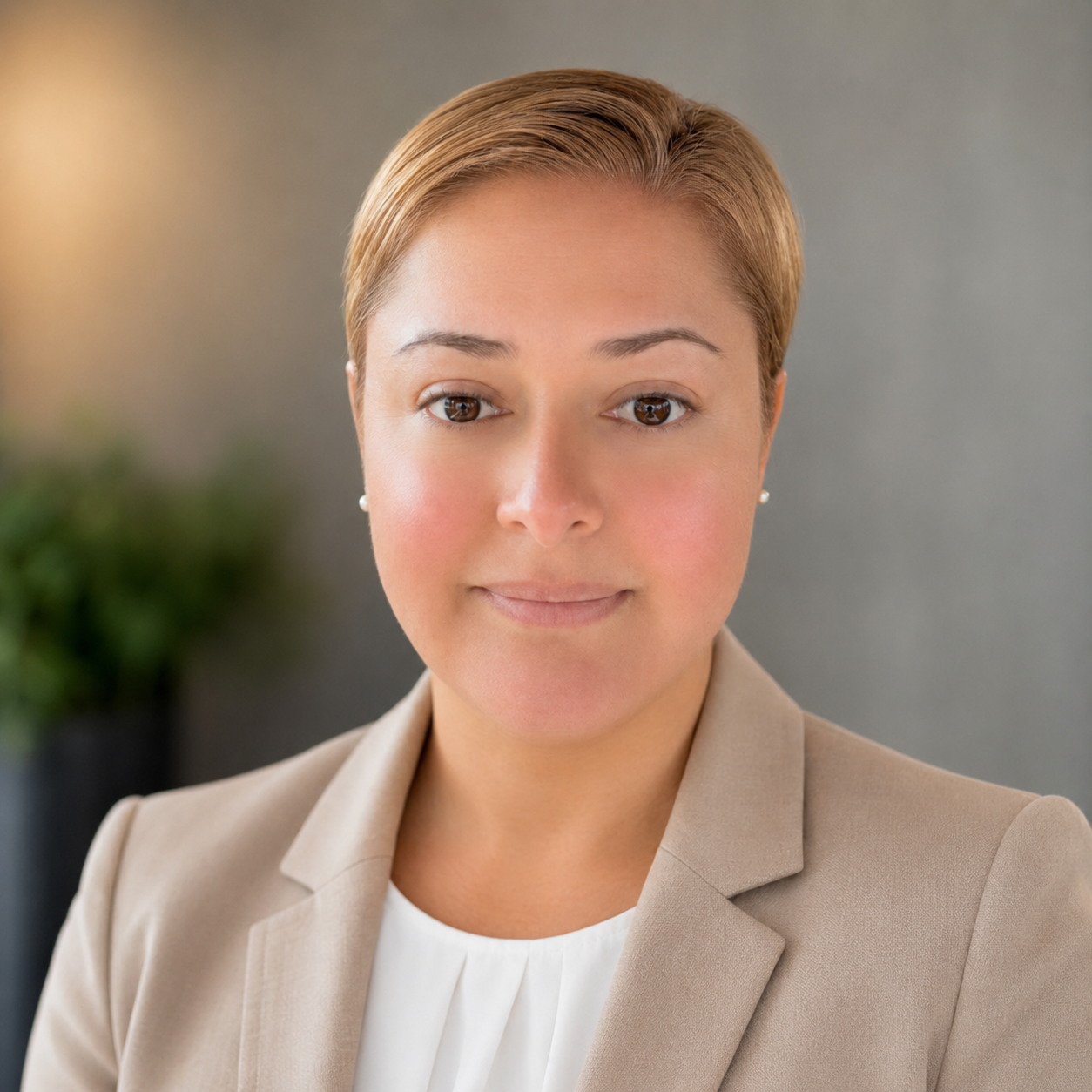 A professional woman with short light brown hair, wearing a beige blazer and white top, looking at the camera indoors.