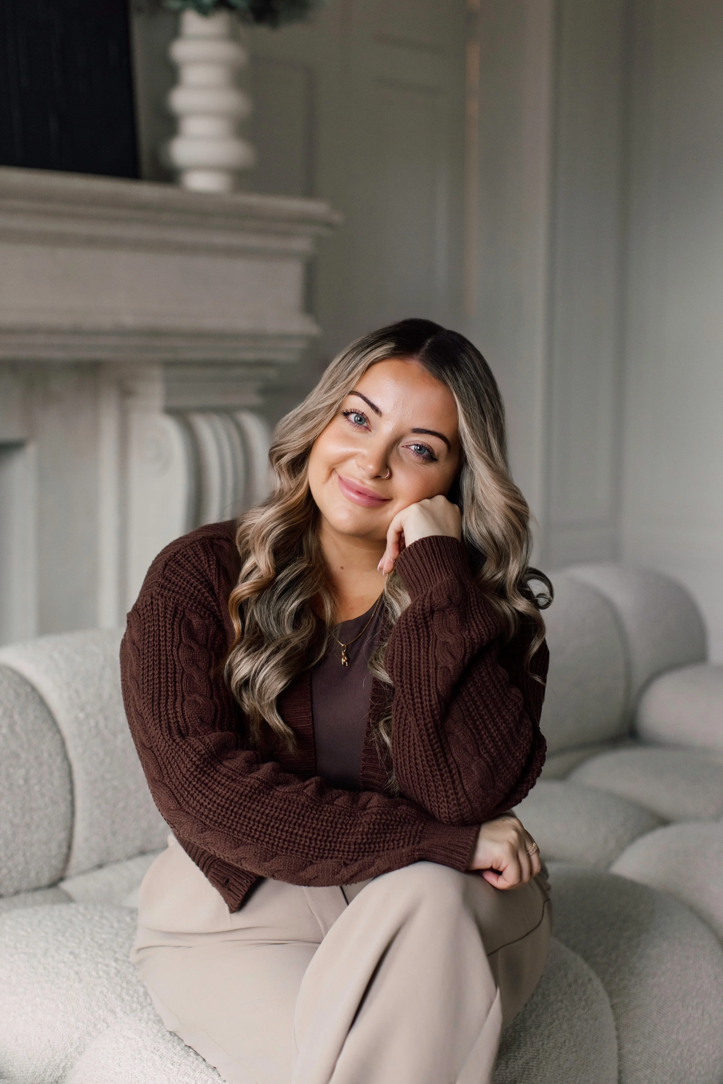Julia Mazzotta, Psychotherapist in Richmond Hill, ON, with long, wavy hair, sitting on a light-colored sofa indoors, wearing a dark brown sweater and beige pants, smiling and resting her face on her hand.