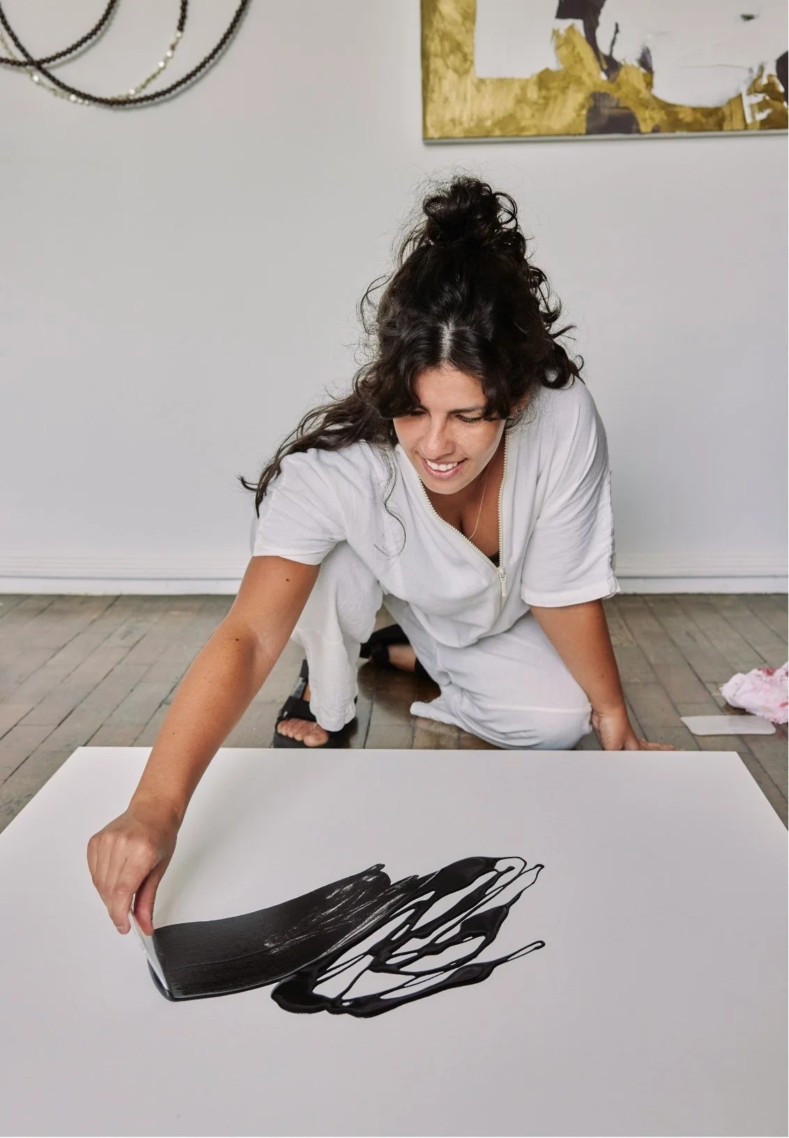 Woman creating a black abstract painting on a white canvas, kneeling on the floor, in a room with minimal decor.
