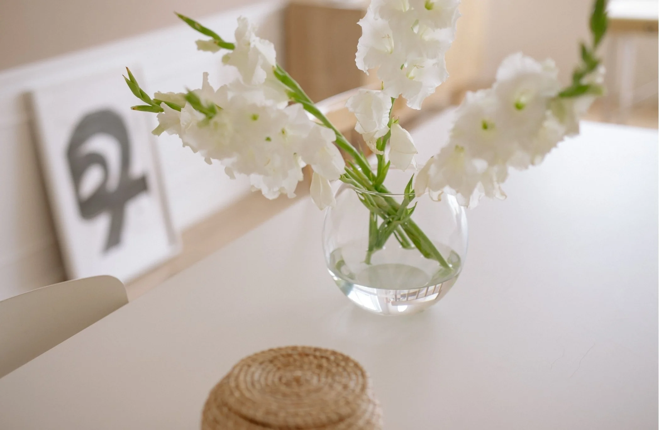 A clear glass vase with white gladiolus flowers on a white table, with woven coasters in the foreground and a blurred background.