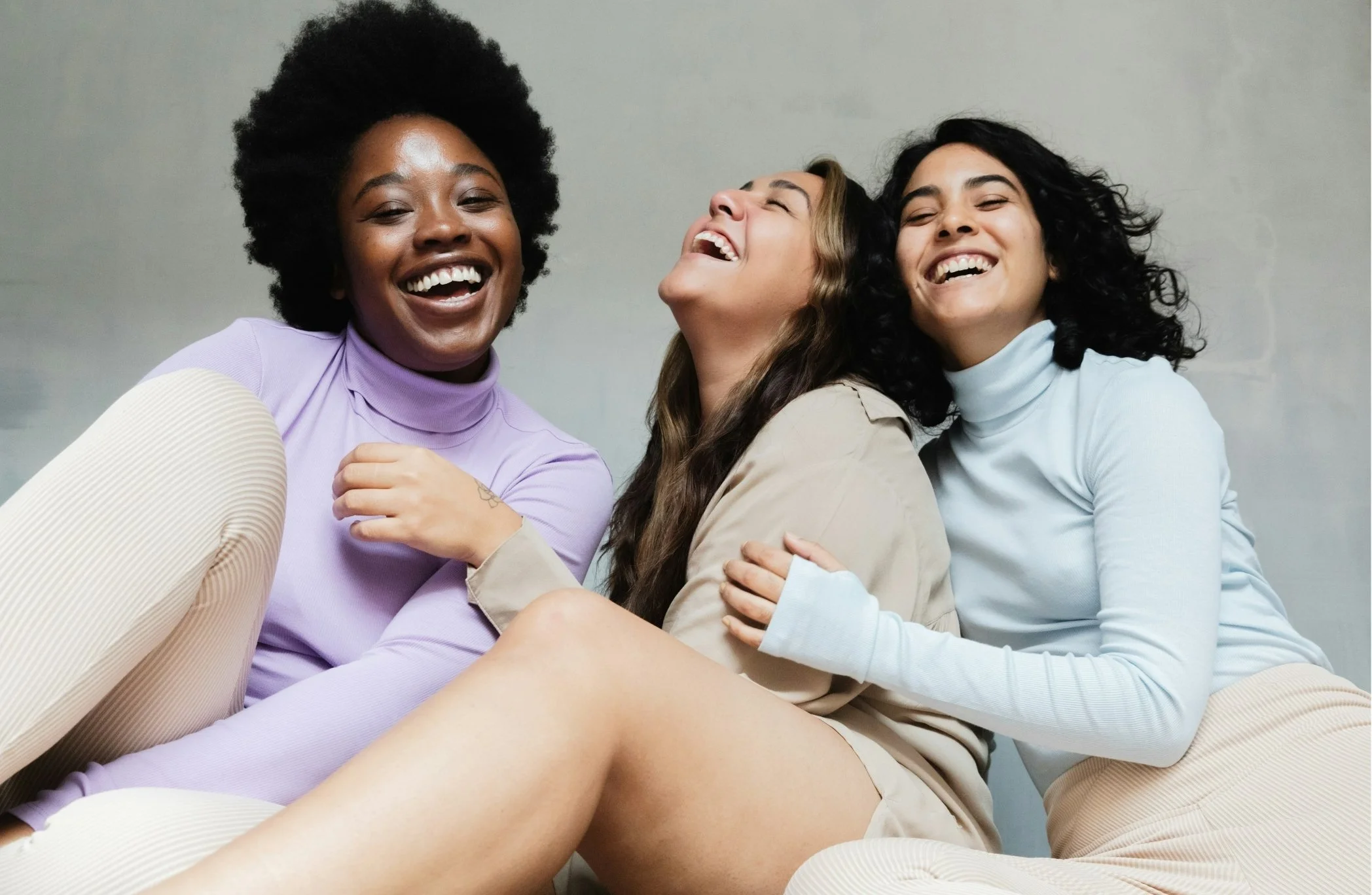 Three women laughing and hugging each other in casual clothing, against a plain gray background.