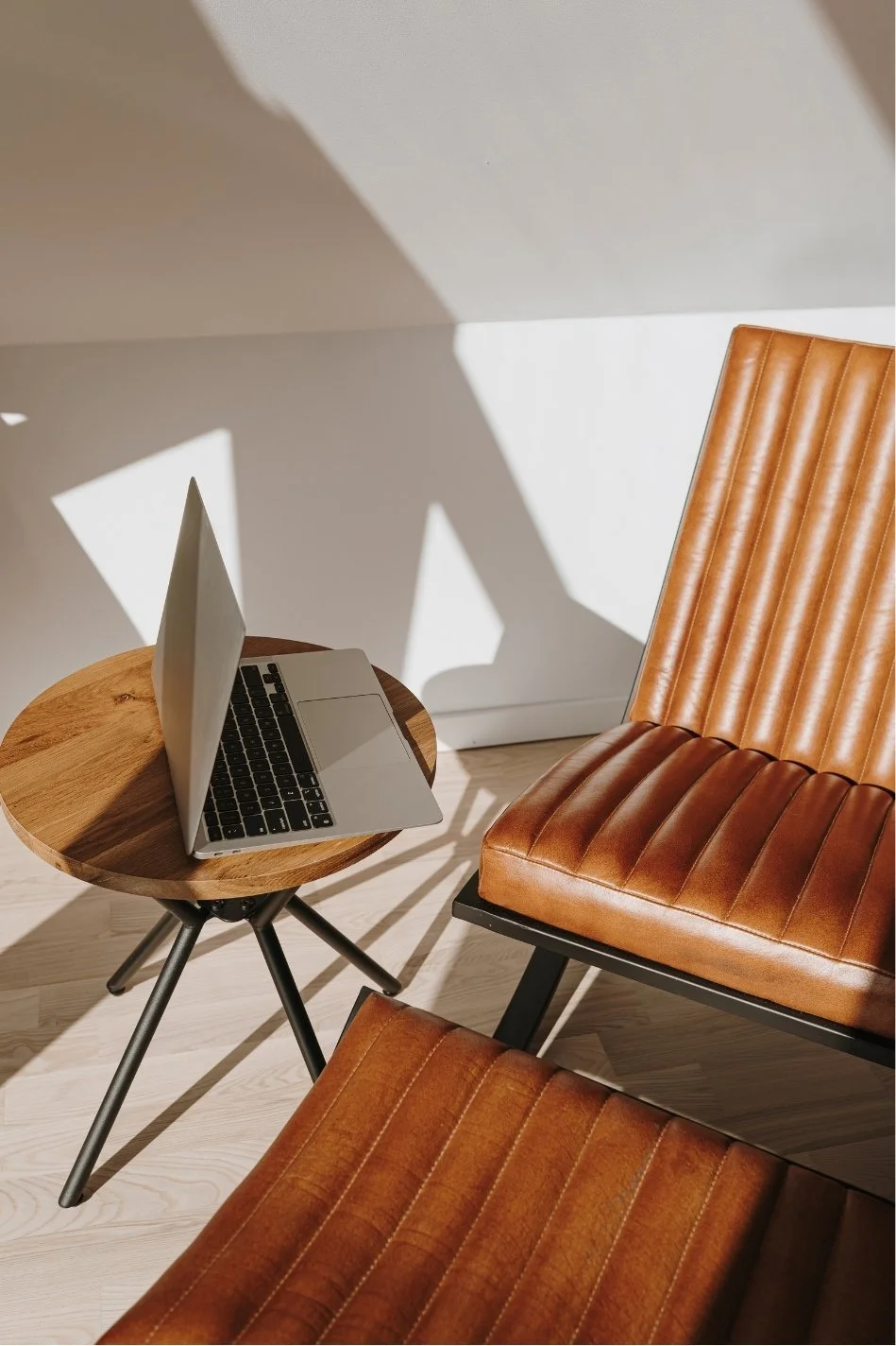 A modern workspace with a small round wooden table, a silver laptop, and a brown leather chair with vertical stitching, illuminated by sunlight creating shadows on the wall and floor.