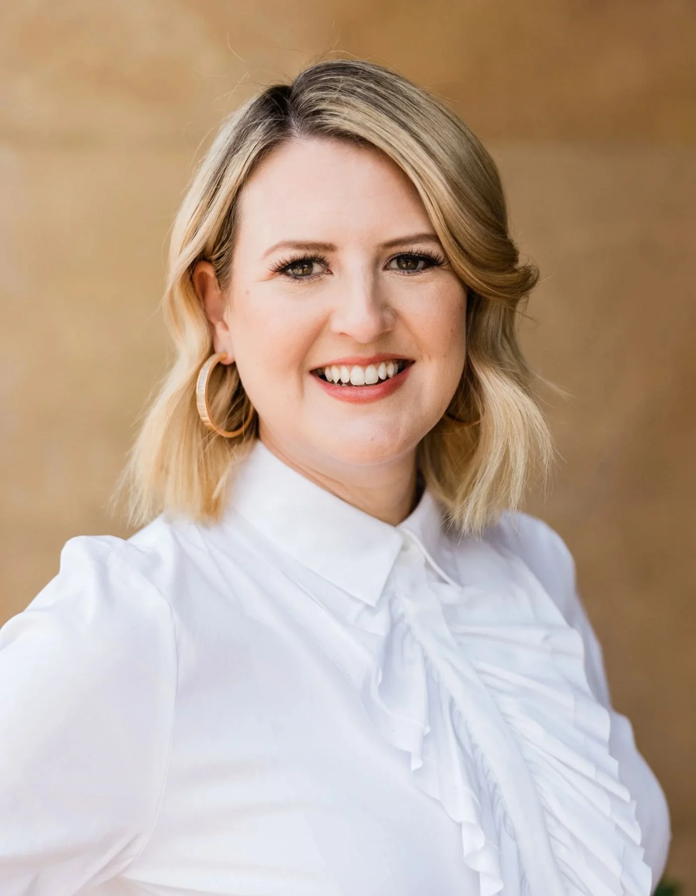 A woman smiling, wearing a white blouse and gold hoop earrings, with a blurred warm-toned background.