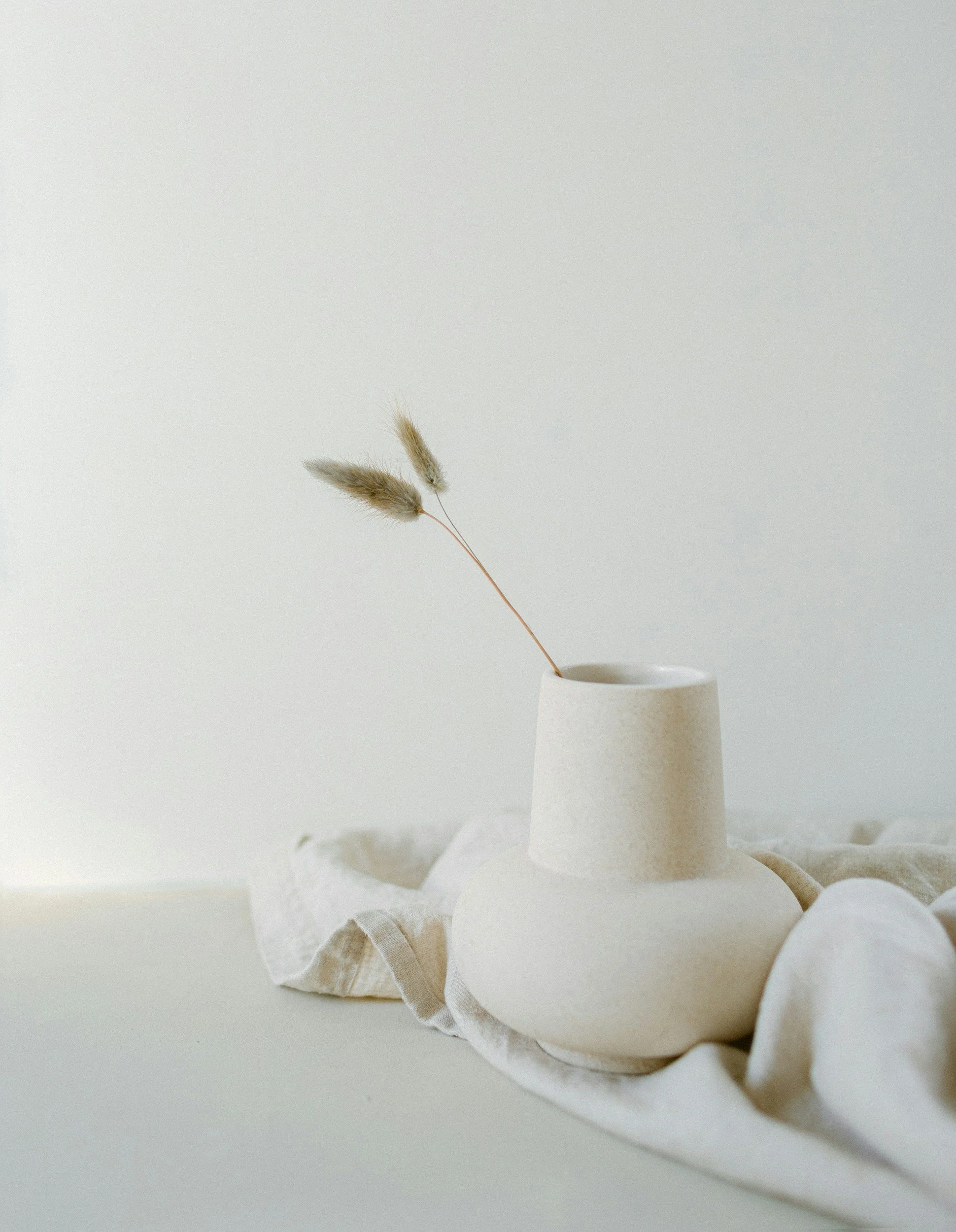 A simple white ceramic vase with dried flowers on a white surface, surrounded by crumpled white fabric.