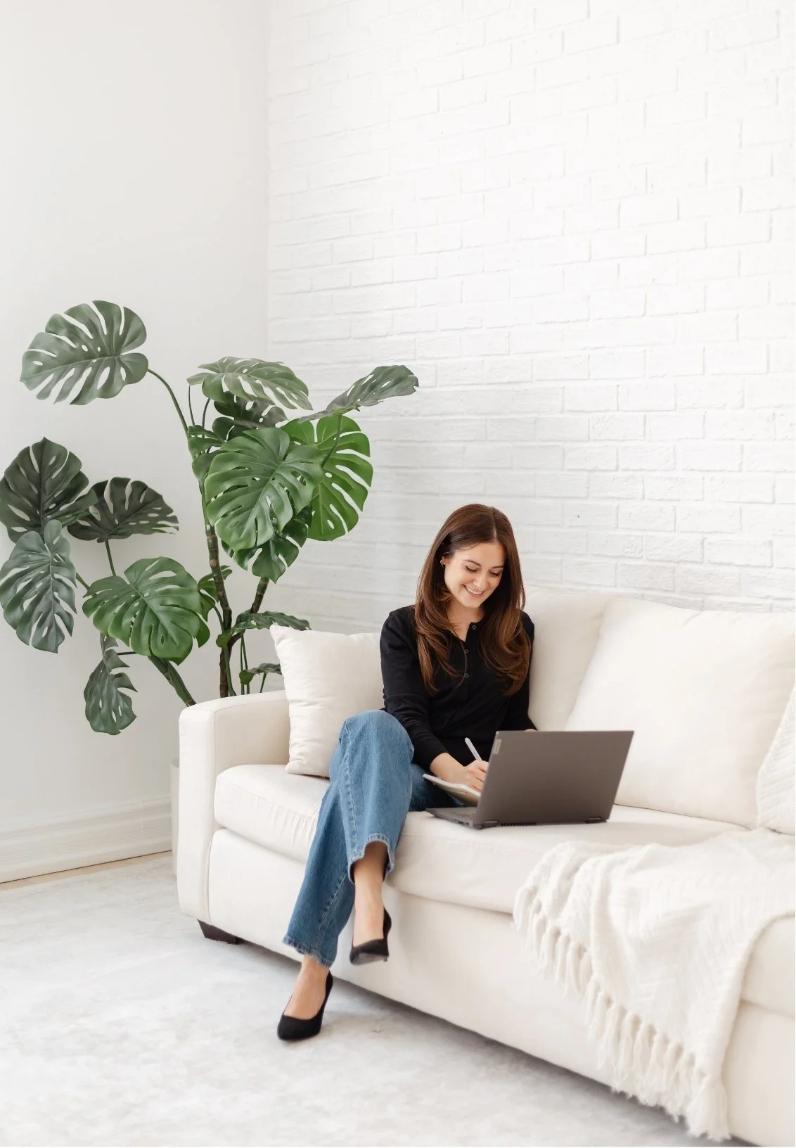 A woman sitting on a white sofa, using a laptop and writing in a notebook, with a large green leafy plant beside her in a bright, modern room with white brick walls.