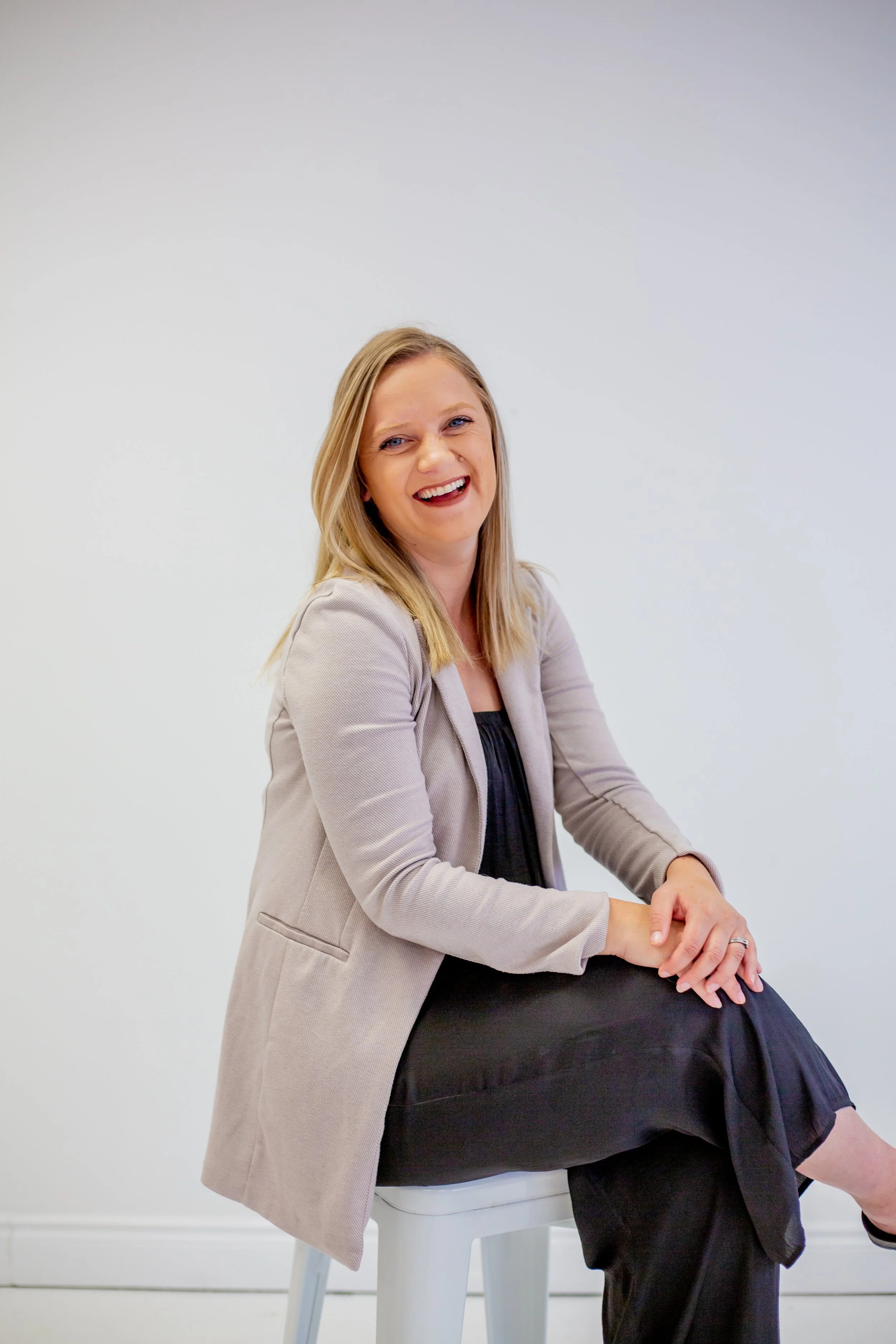 Sara Boersma, Psychotherapist,  in Richmond Hill, ON, with blonde hair sitting on a white stool in front of a plain white wall. She is wearing a light-colored blazer over a black top and smiling.