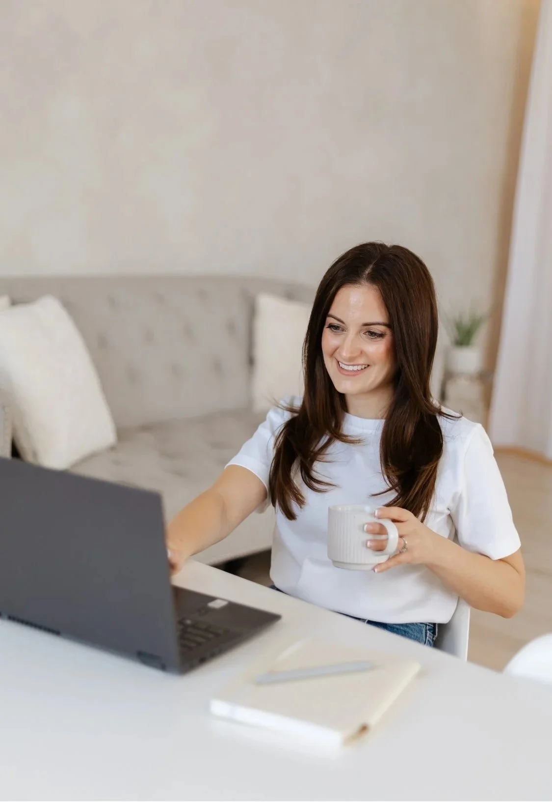 Samantha Mirarchi, therapist in Richmond Hill, ON, with long brown hair sitting at a desk, smiling, holding a white coffee mug, using a laptop in a bright room with a beige couch and a small plant in the background.