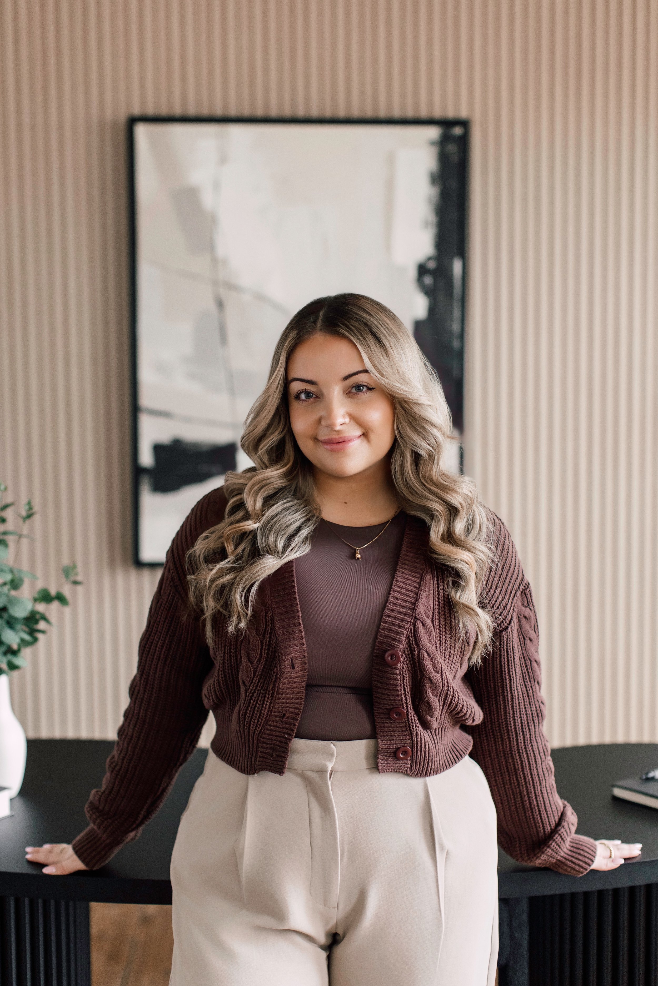 Julia Mazzotta, Psychotherapist in Richmond Hill, ON, with long, wavy blonde hair standing at a black table in a room with a black and white abstract painting in the background. She is wearing a brown cardigan and matching top with cream pants.