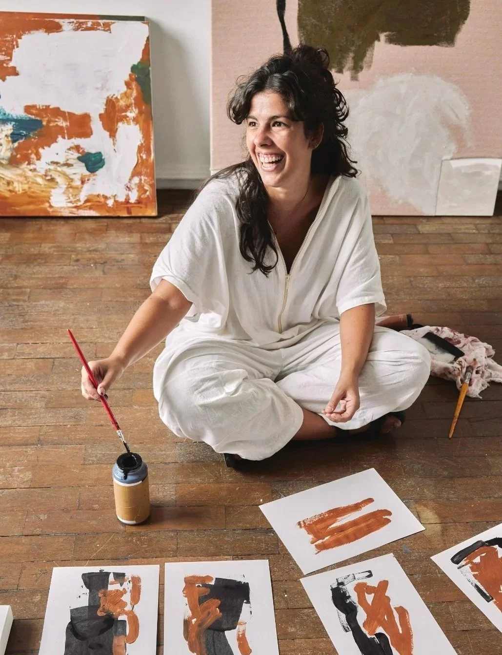 A woman sitting cross-legged on a wooden floor, smiling, holding a brush dipped in black paint. Behind her are abstract paintings with bold brushstrokes. Several sheets of paper with abstract artwork are in front of her.