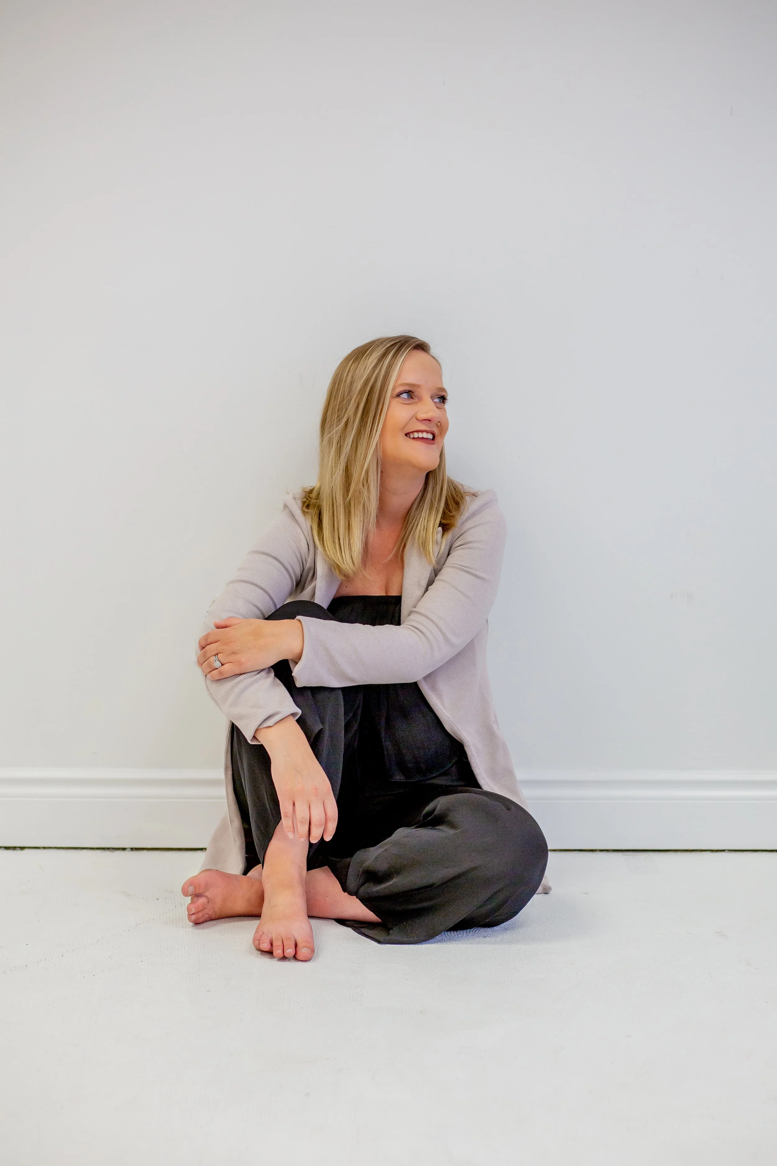 Sara Boersma, Psychotherapist,  in Richmond Hill, ON, with blonde hair sitting on the floor against a white wall, smiling and looking to her right, with her legs crossed and arms resting on her knees.